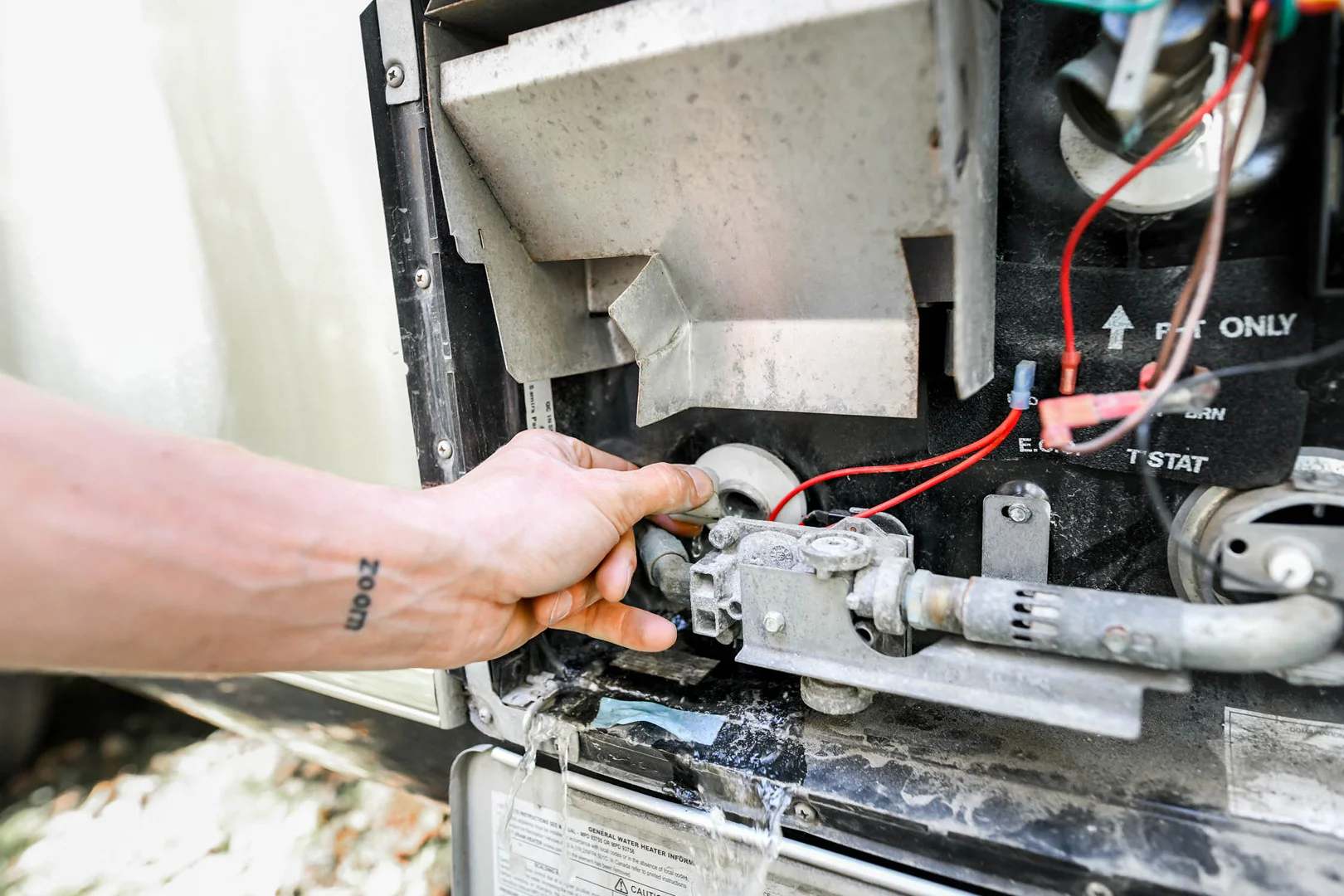 A person’s hand is adjusting a component inside an open water heater compartment, with water leaking from a pipe below and wires visible near the panel.