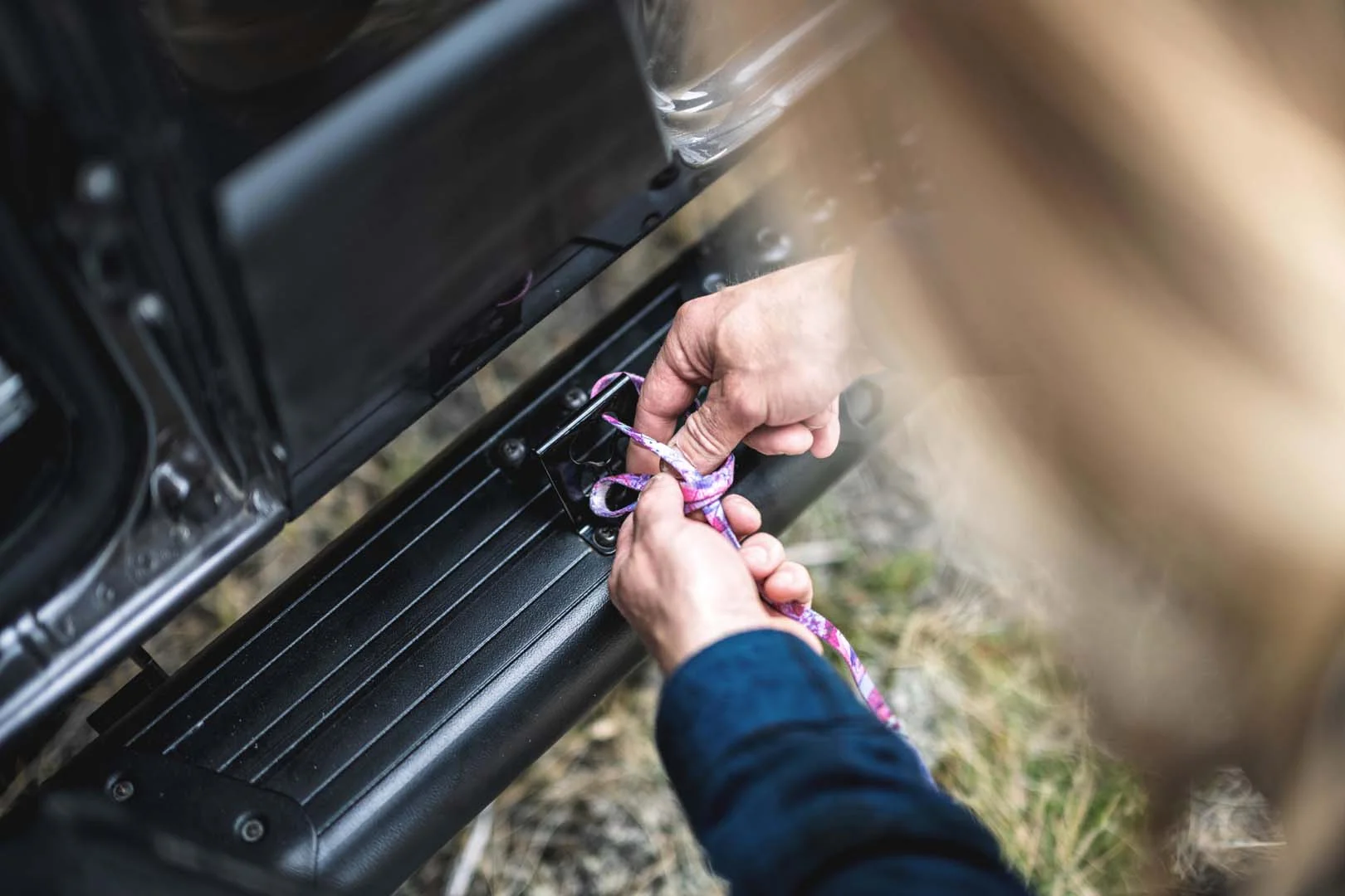 Hands tying a rope to a hook on a vehicle's side step, close-up view, with grass visible in the background.