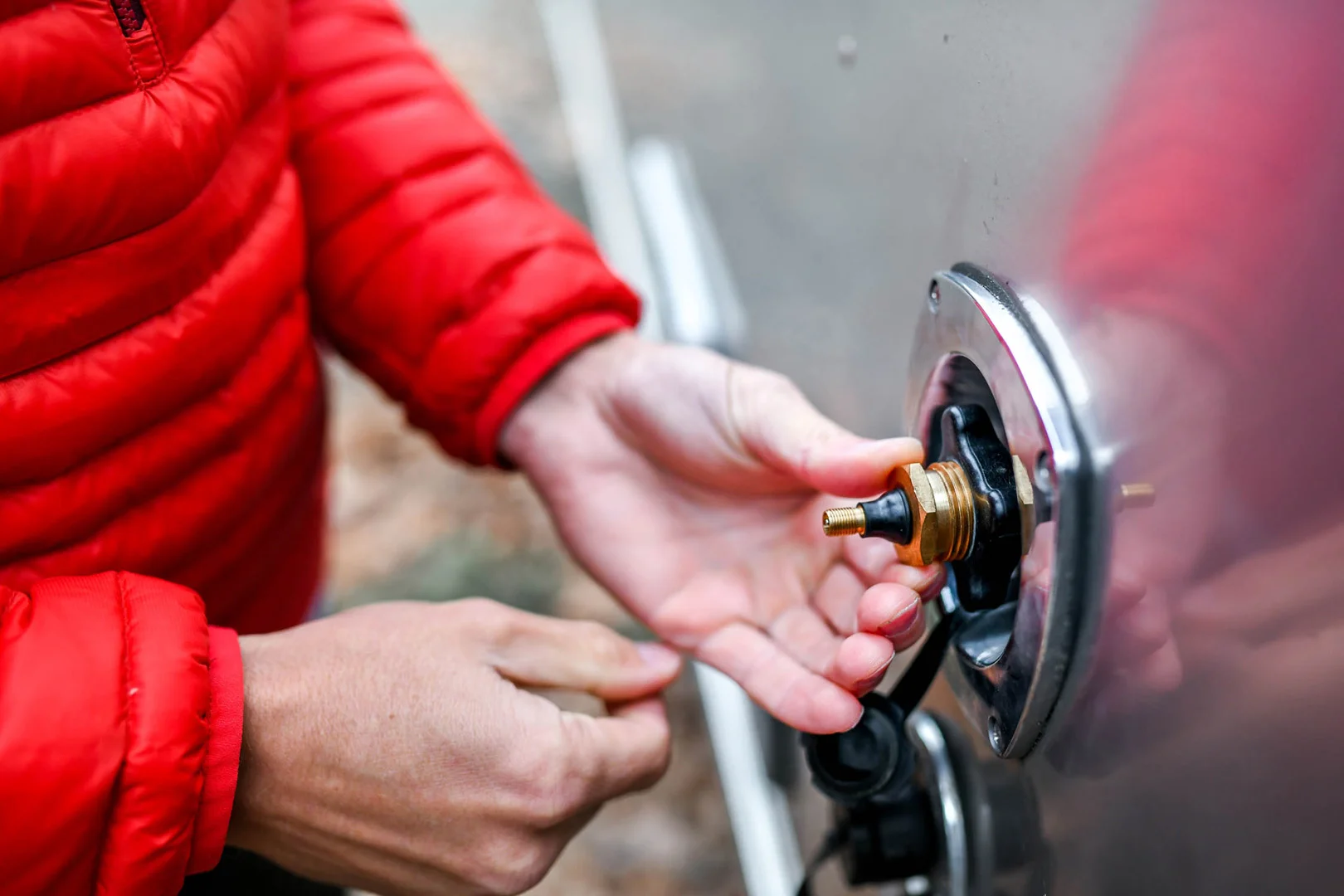 A person in a red jacket uses both hands to install or replace a valve stem on a shiny metal surface, possibly part of a vehicle or equipment, with a blurred outdoor background.