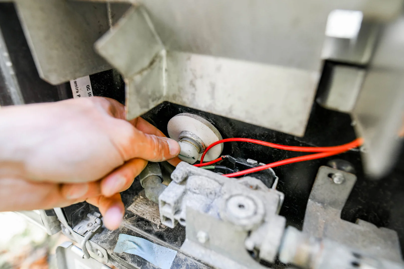A close-up of a persons hand adjusting a component inside a mechanical device with visible red electrical wires and metal parts, suggesting repair or maintenance work.