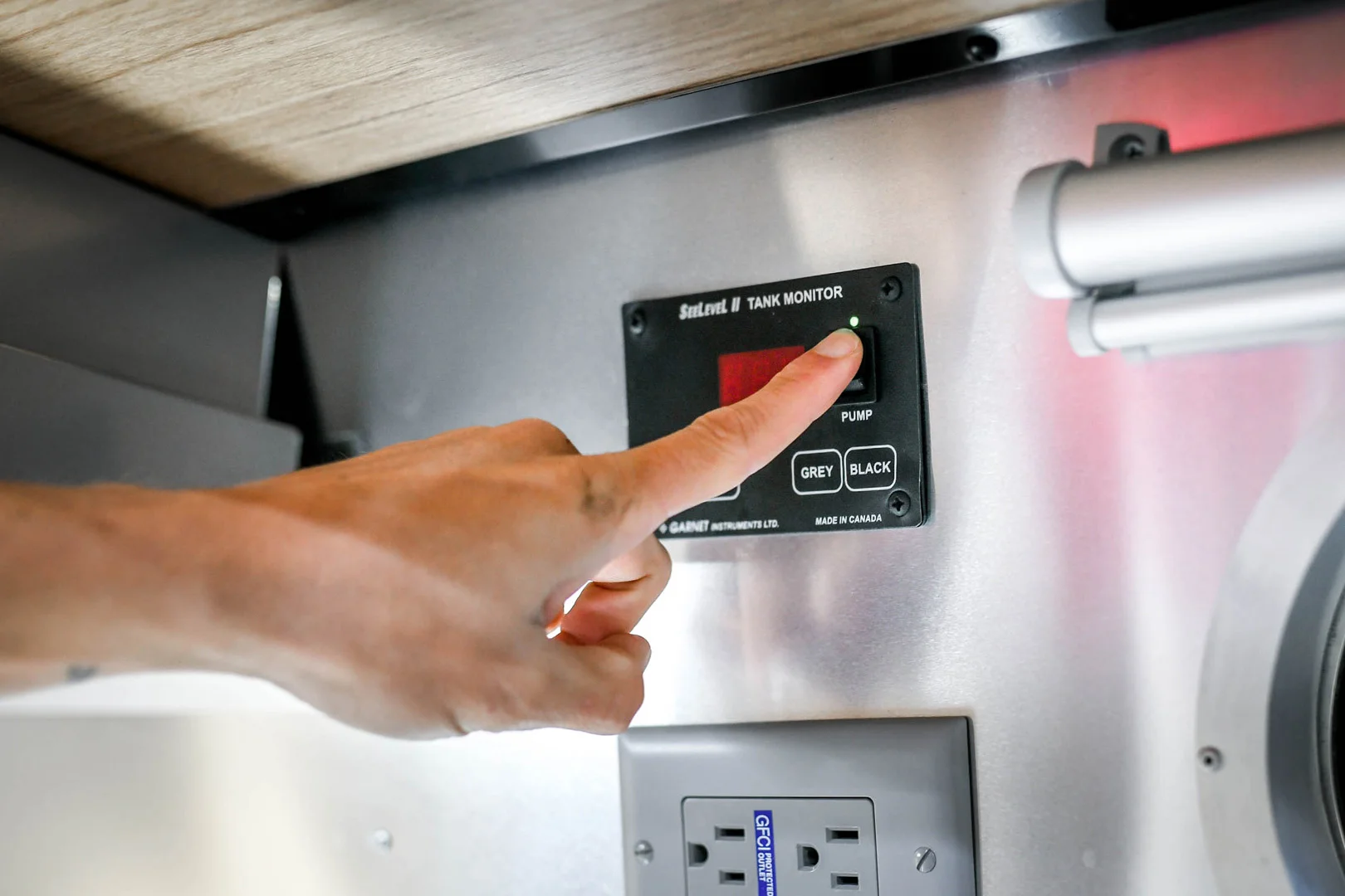 A hand presses a button on a black tank monitor panel labeled SaniConX above a wall outlet, with options for Pump, Grey, and Black tanks inside a metallic enclosure.
