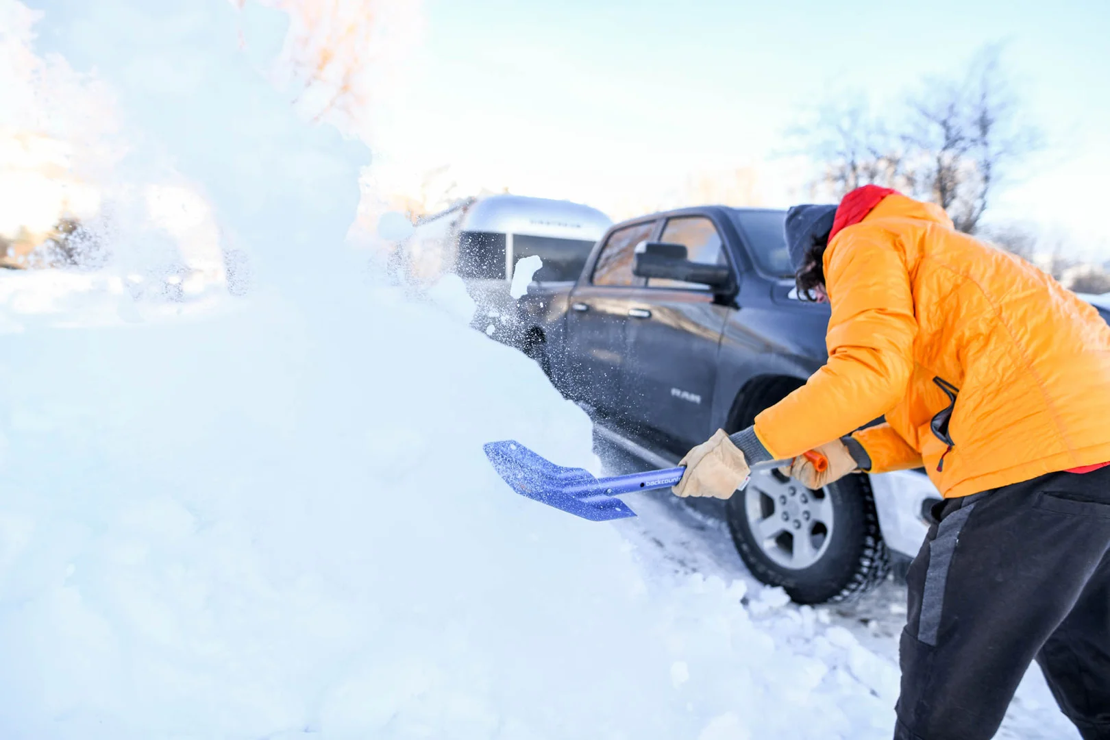 Person in an orange jacket shoveling snow next to a parked black truck on a clear day.