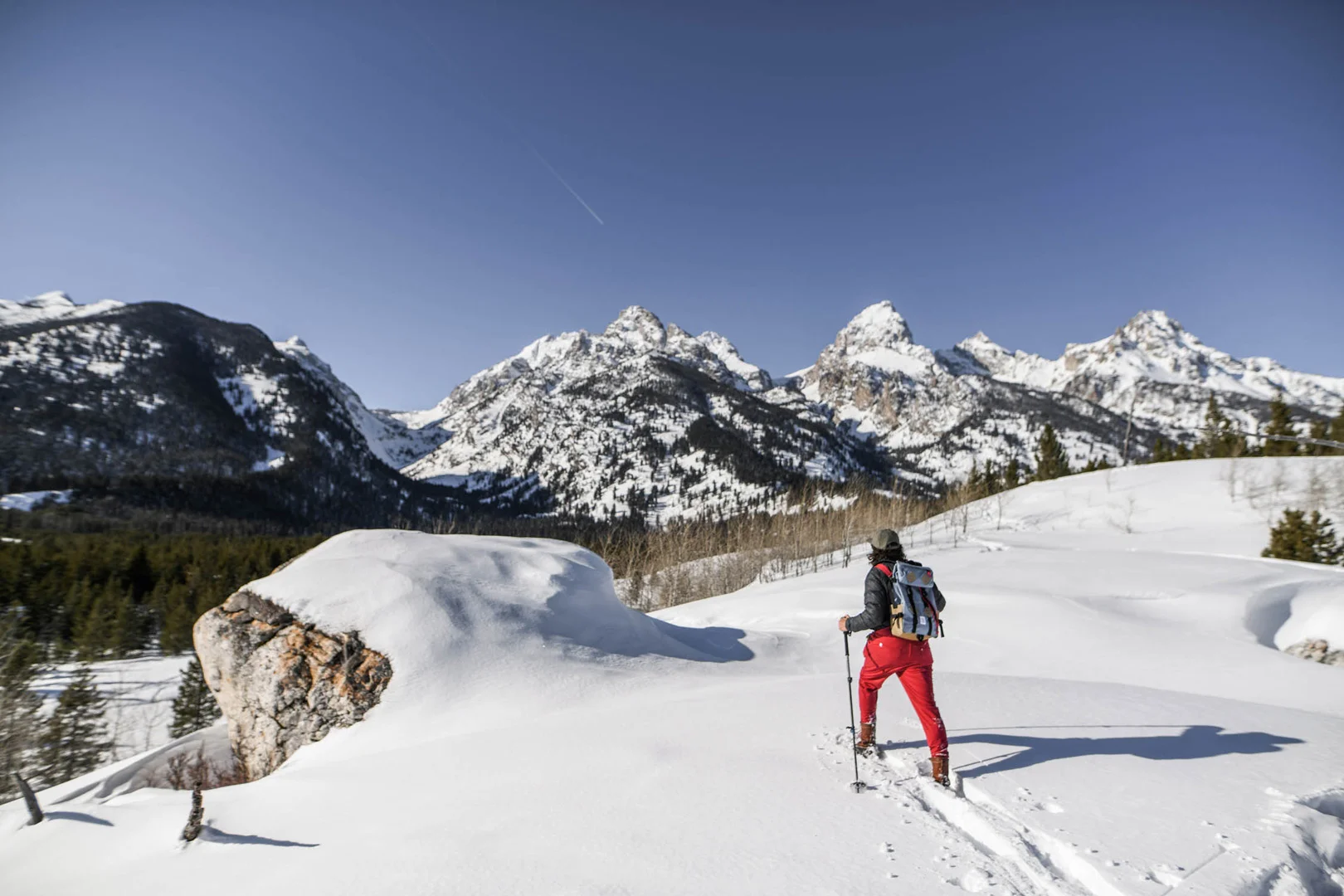 A person in red pants snowshoeing toward a snowy mountain range under a clear blue sky.