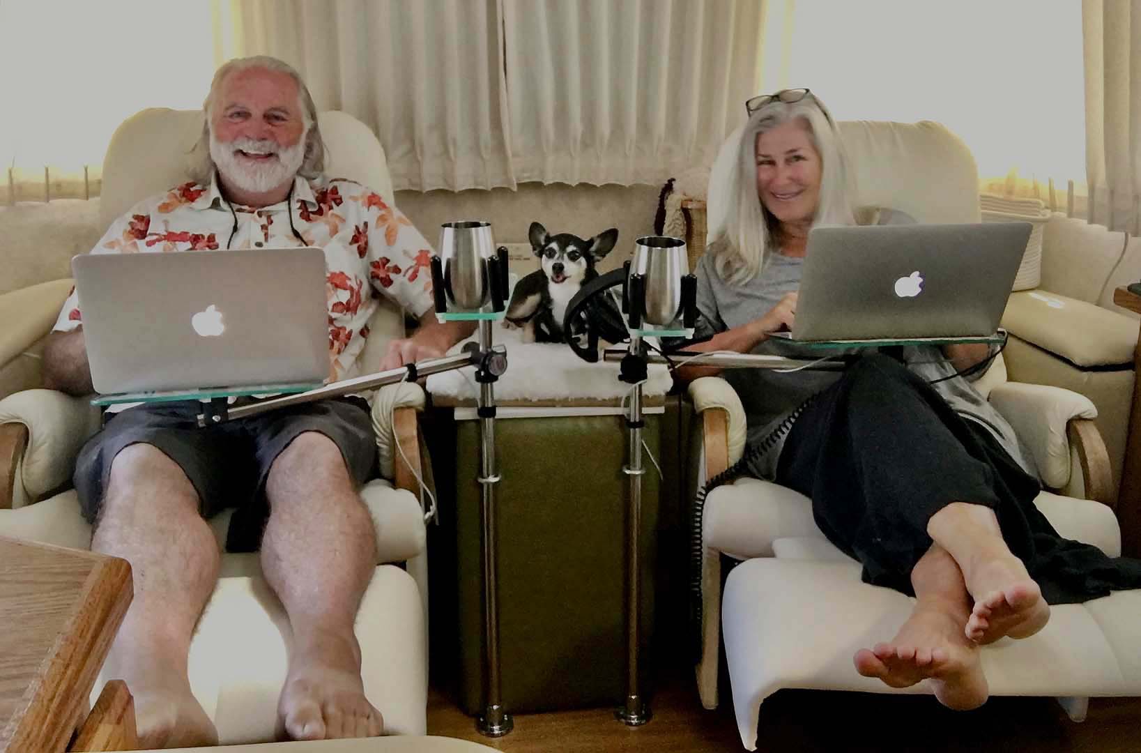 A man and woman sit comfortably on recliners with laptops on their laps, a small black and white dog between them, in a cozy, warmly lit room.