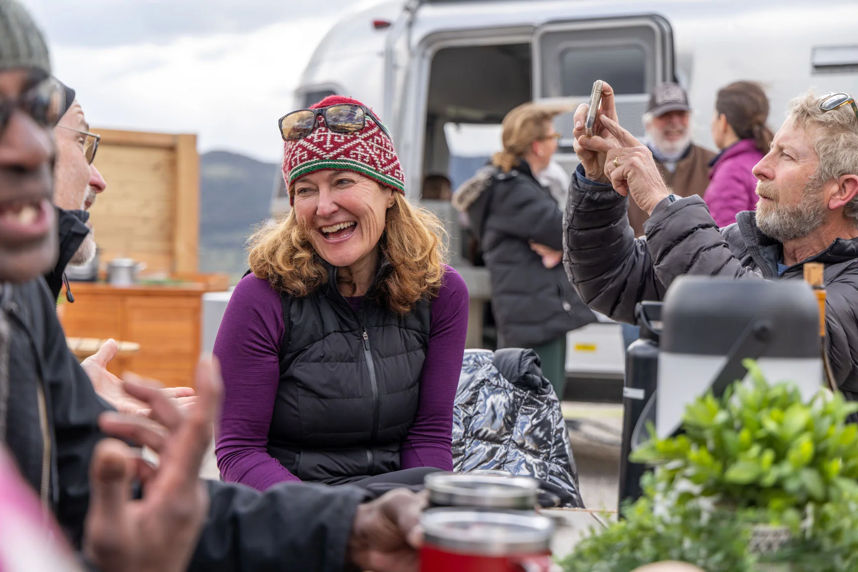 Group enjoying an outdoor gathering by a camper, with a woman in a knit hat laughing and a man taking photos on a phone.