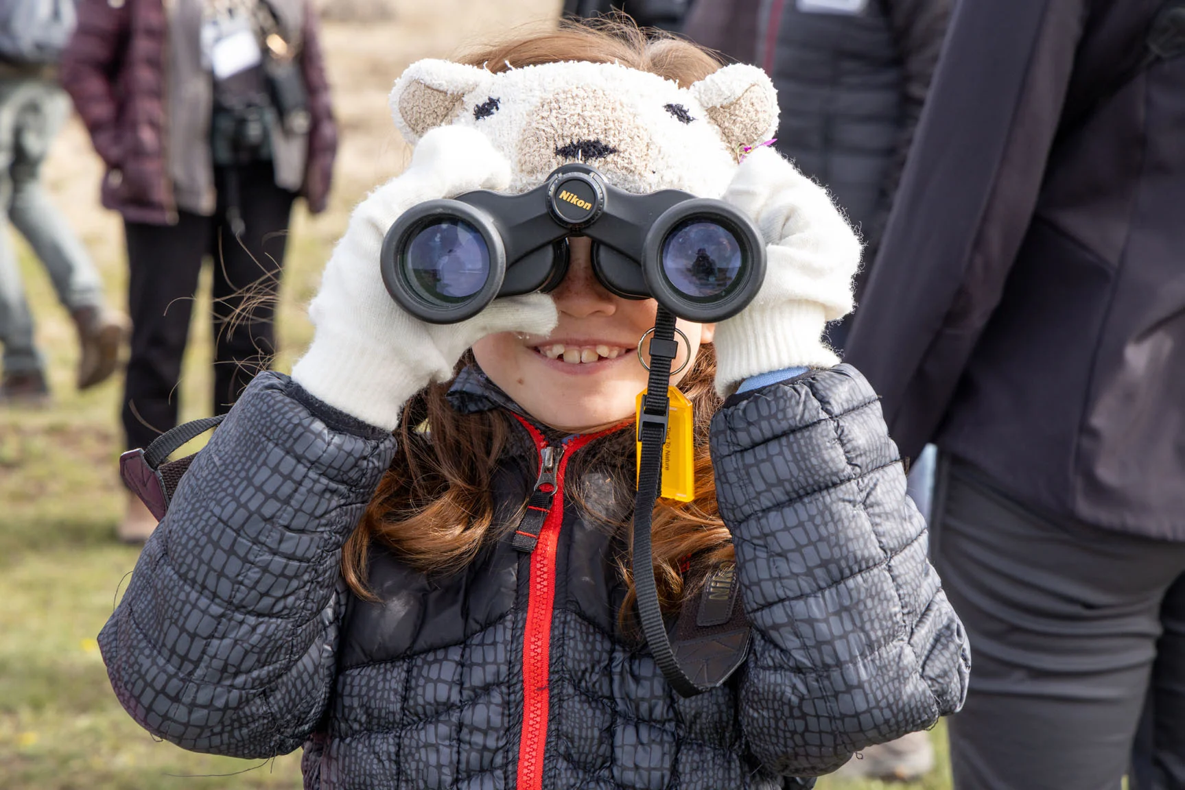 Child wearing a bear hat and gloves, smiling while looking through binoculars outside.