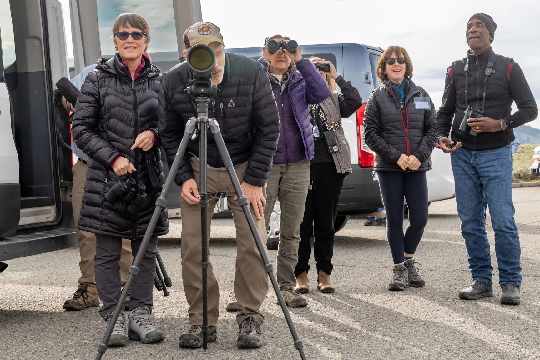 A group of people engaged in birdwatching, standing outdoors with binoculars and a telescope, next to parked vehicles.