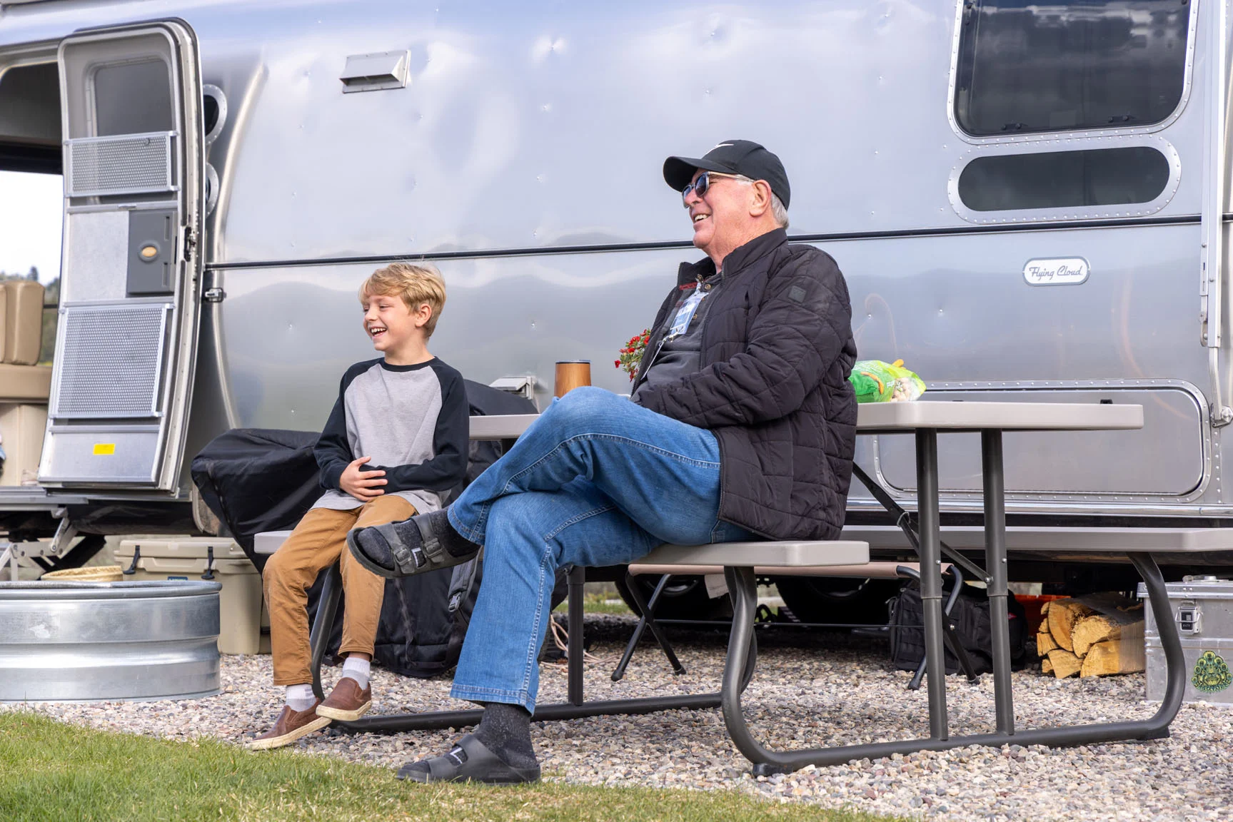 A man and a child sit on a picnic table outside an Airstream trailer, smiling and enjoying the outdoors. The man wears a cap and jacket.
