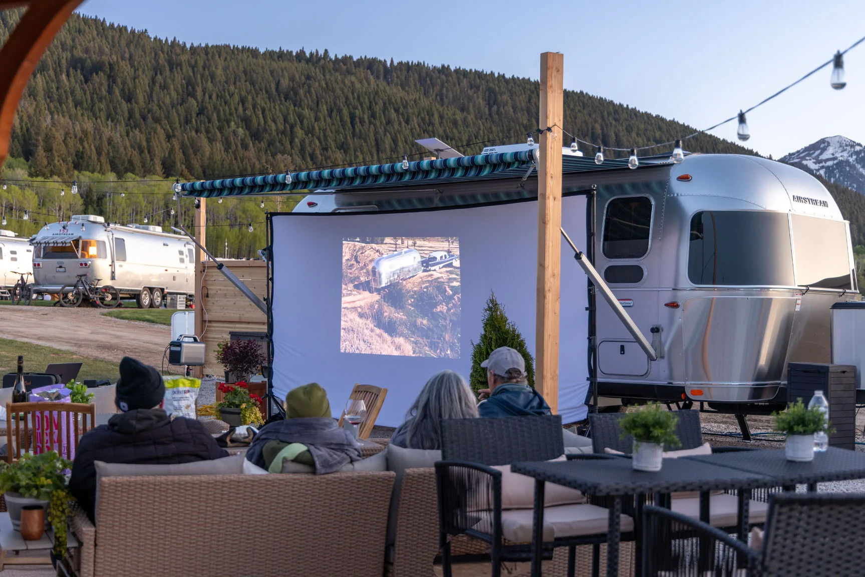 People watch a movie on an outdoor screen next to a silver RV in a scenic campsite with mountains in the background.