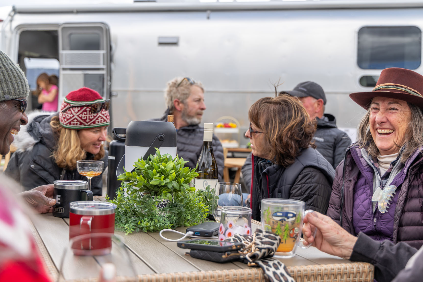 A group of people, bundled in winter clothes, laugh and chat around a table outdoors, with a large silver trailer in the background.