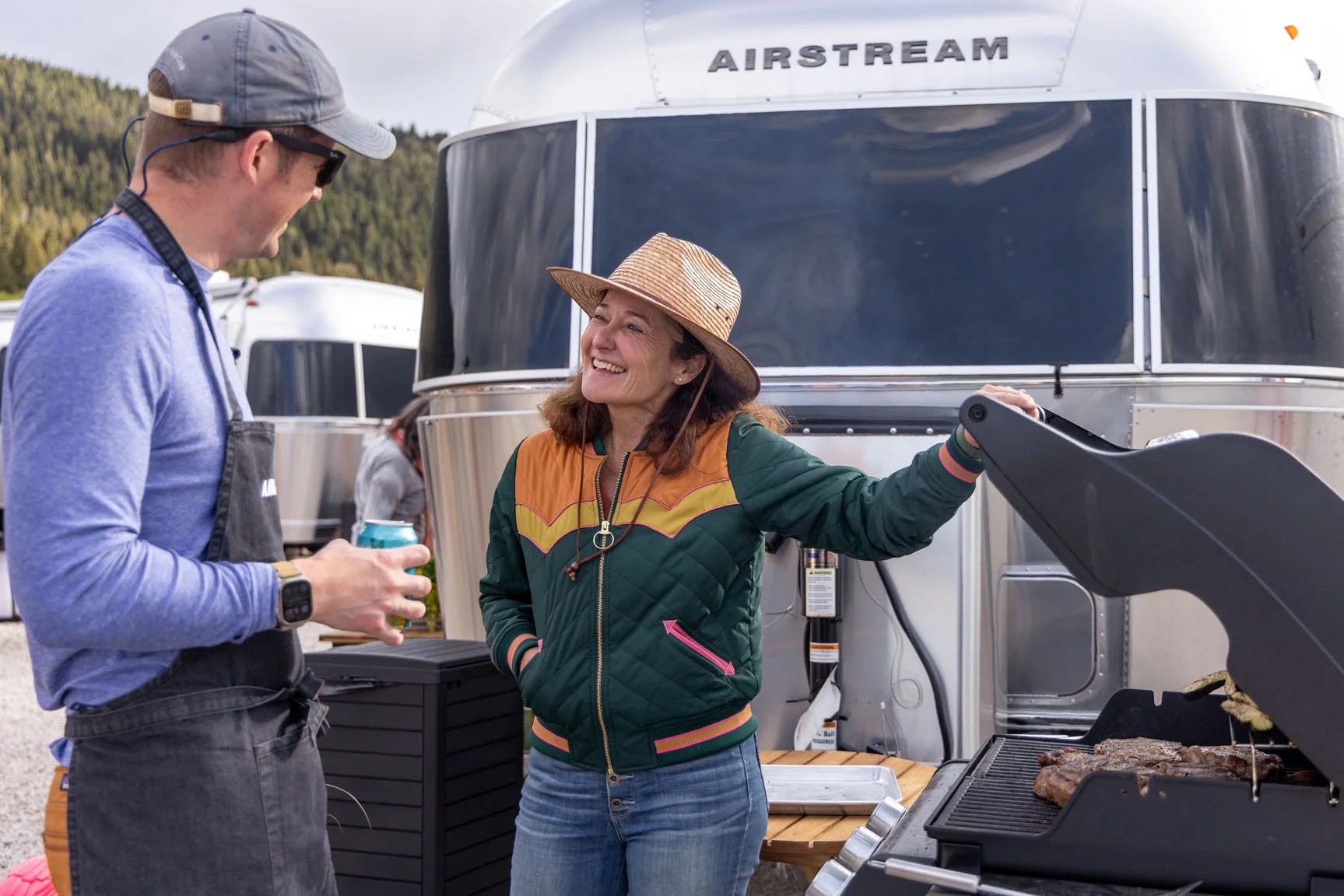 Two people conversing happily beside a grill with steaks, standing near a parked Airstream trailer in an outdoor setting.