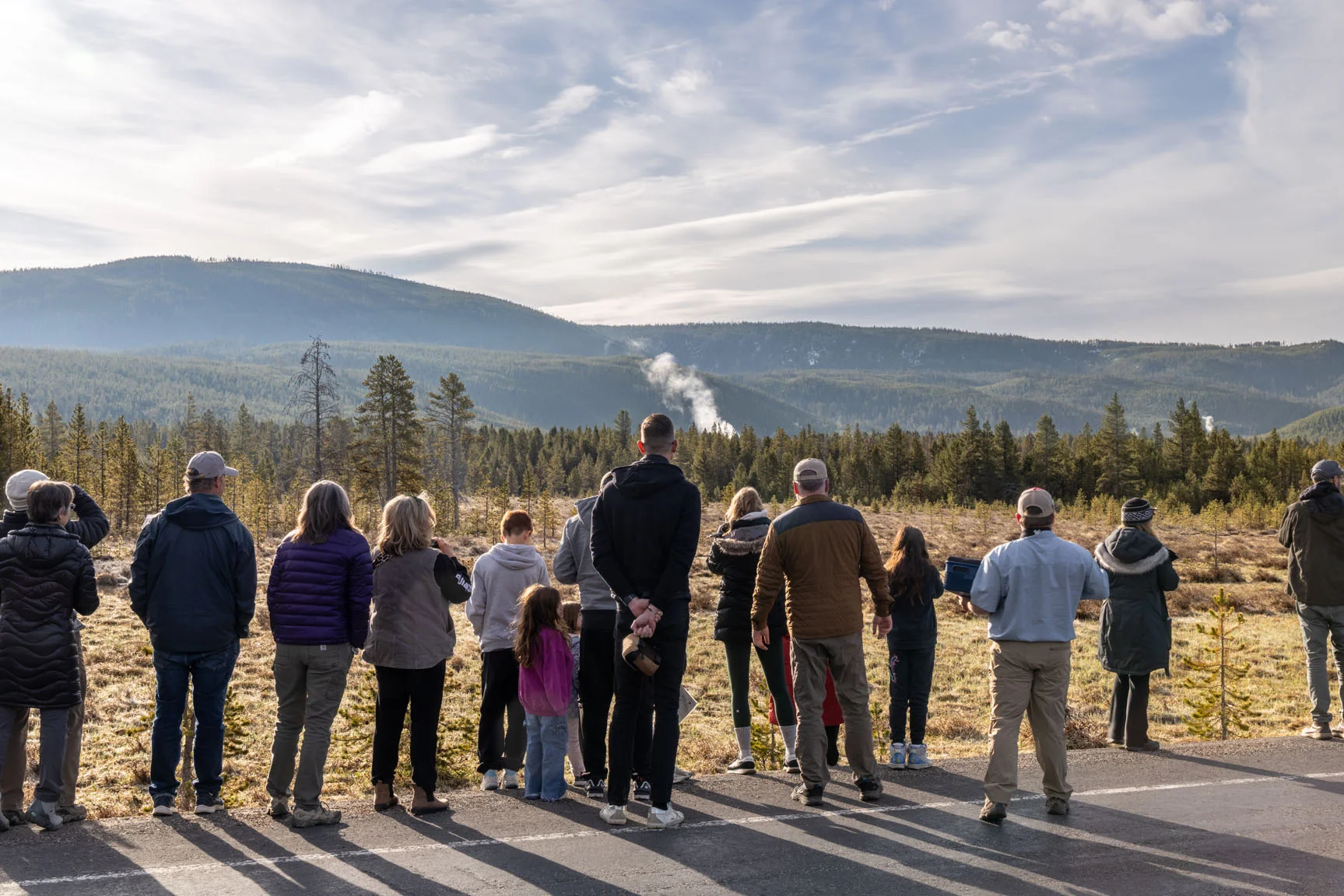 A group of people stands on a path, observing a distant geyser amidst a forested landscape under a cloudy sky.