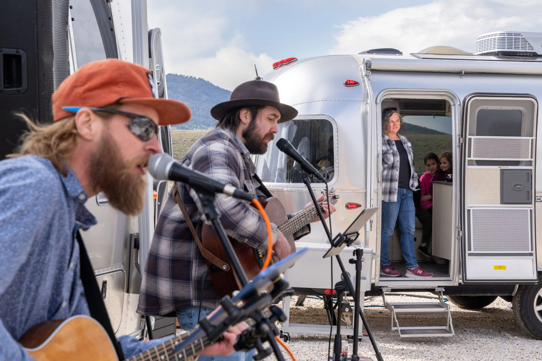Musicians perform outdoors near a silver camper, with a woman and children watching from the doorway. Mountains are visible in the background.