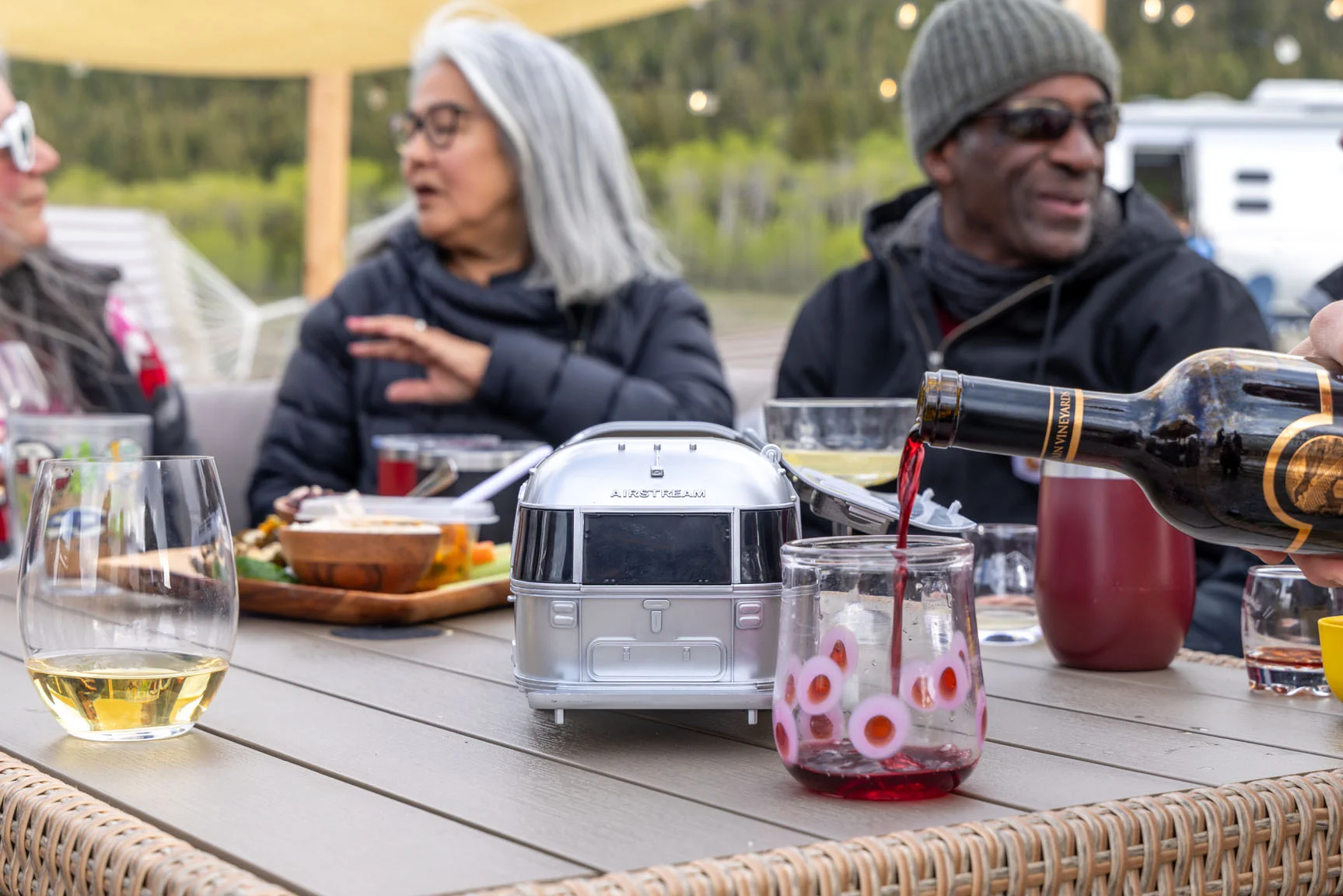 People enjoying a casual outdoor gathering, seated around a table with drinks and snacks, featuring a drink being poured into a glass.