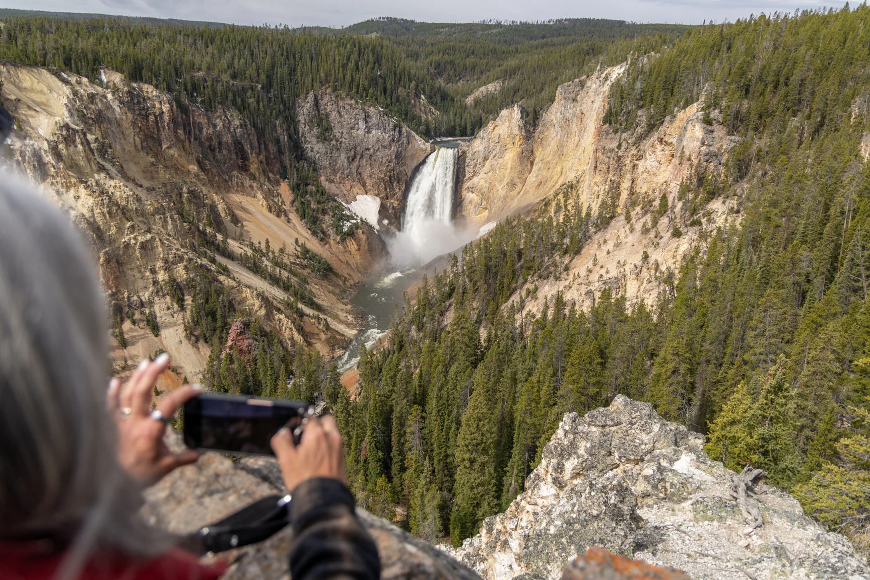 A person takes a photo of a large waterfall cascading into a canyon filled with dense forest under a cloudy sky.