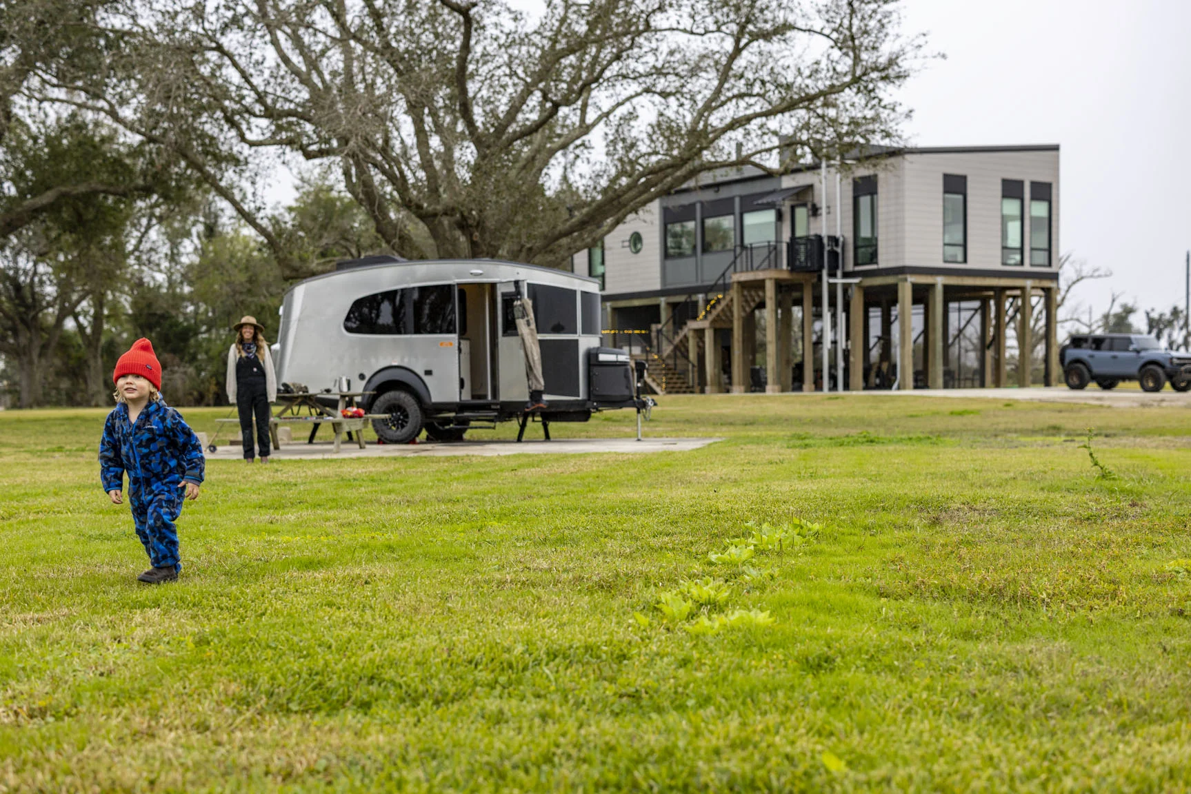 Child in blue outfit and red hat runs on grass near a modern, elevated house and a silver camper trailer; adult stands nearby.