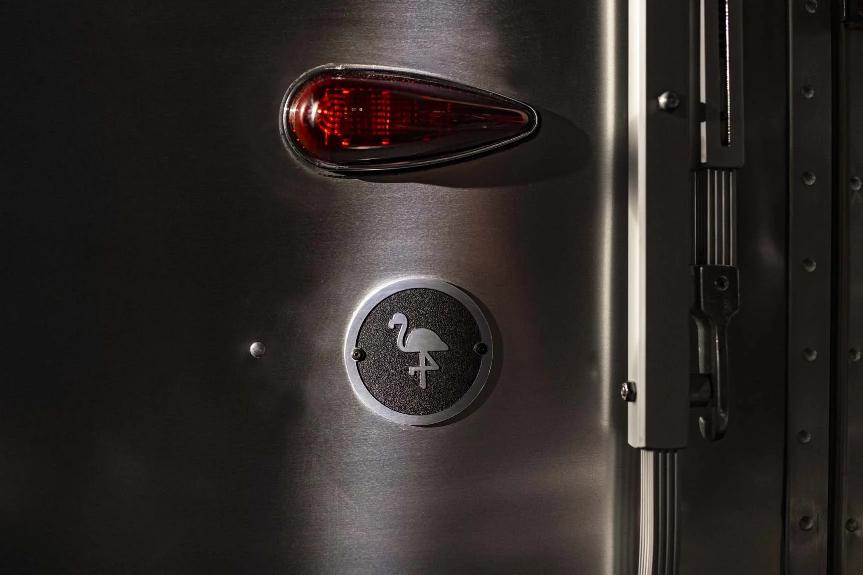 A stainless steel camper with a red teardrop tail light and a circular flamingo emblem against a dark background.
