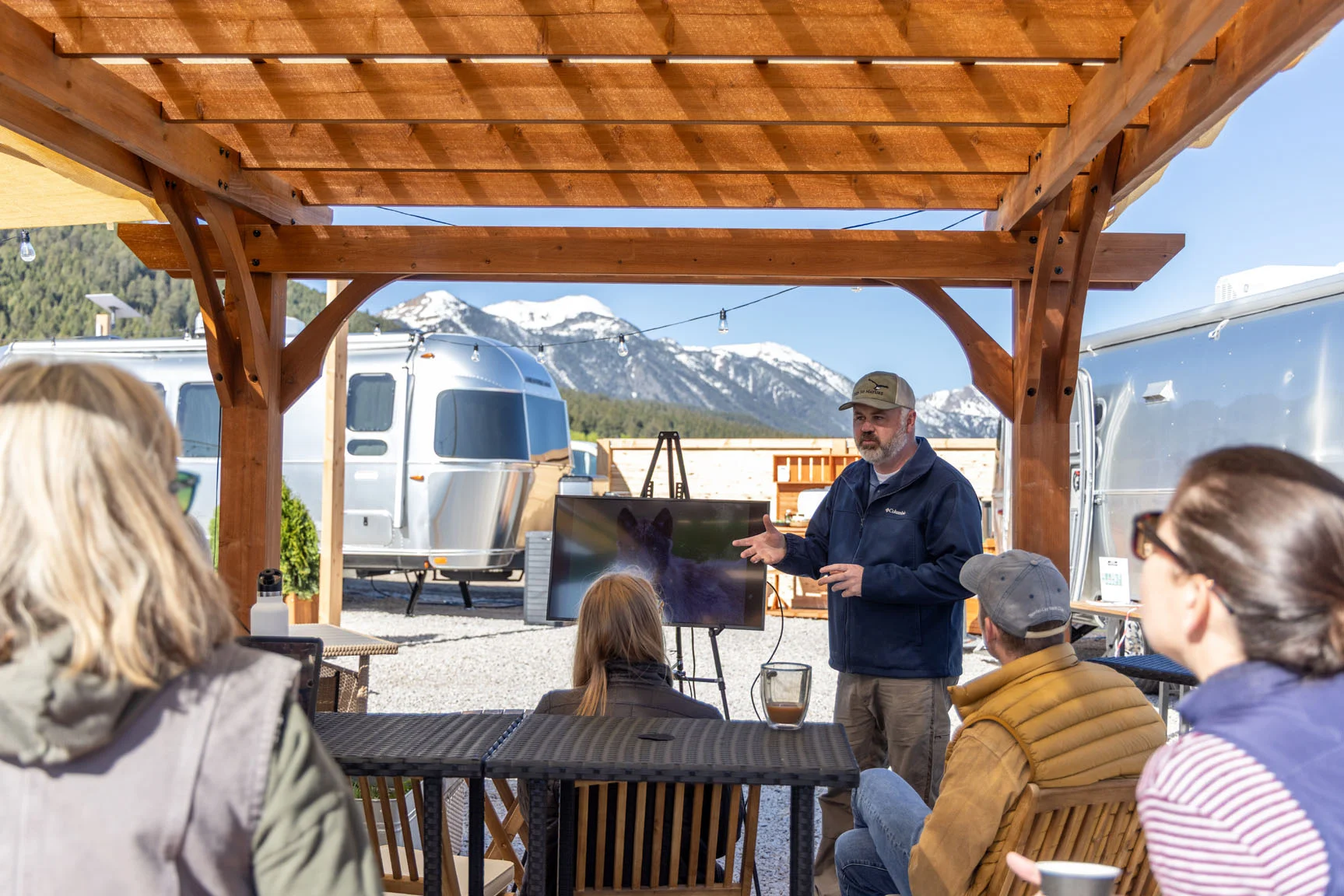 A man gives a presentation under a wooden pergola, with an audience seated and snowy mountains in the background. Campers are visible nearby.