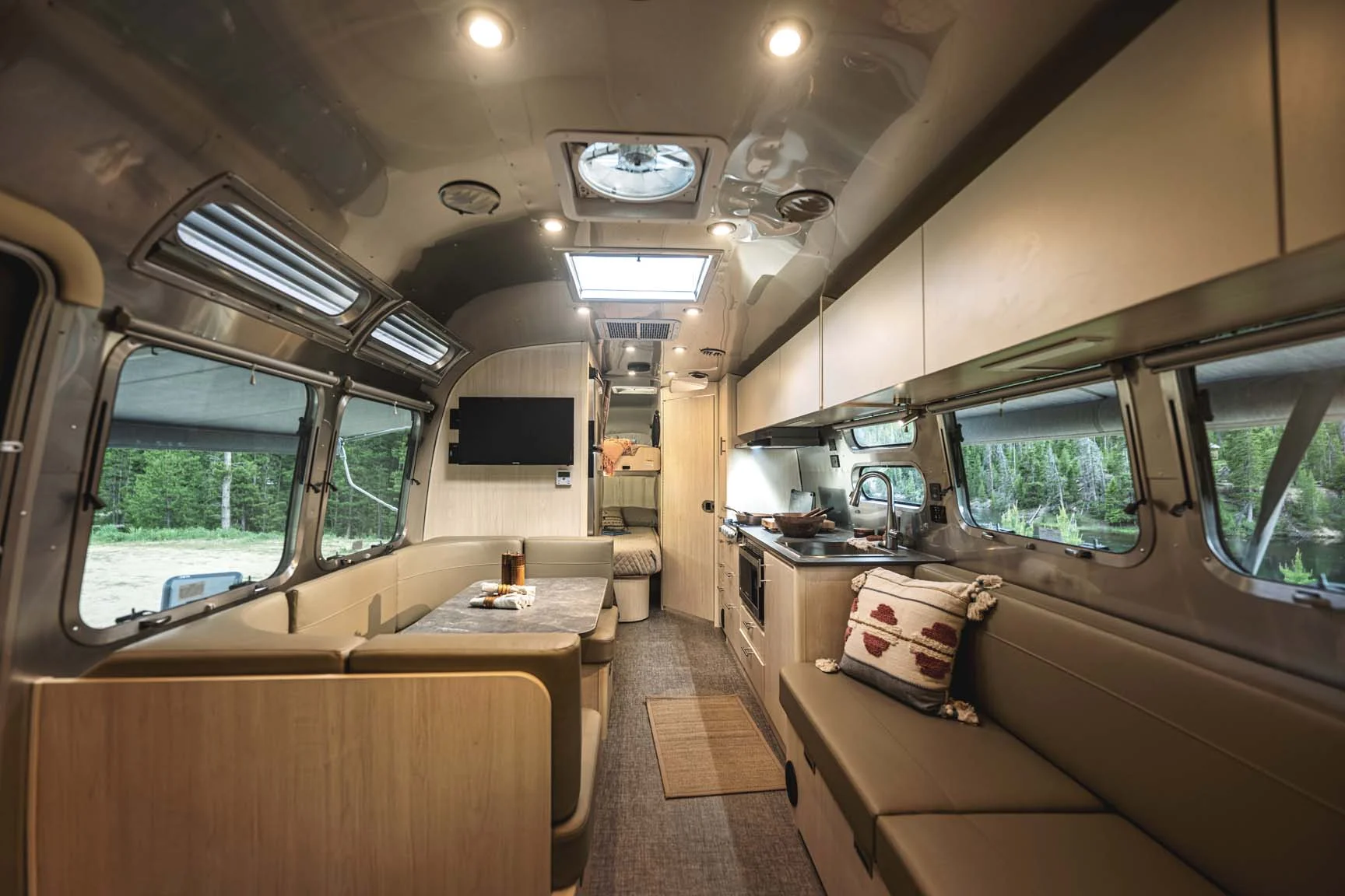 Modern Airstream trailer interior with a dining area, sofa, kitchenette, and sleeping area in the background. Large windows offer views of trees and greenery outside, and the space is well-lit with overhead lighting.