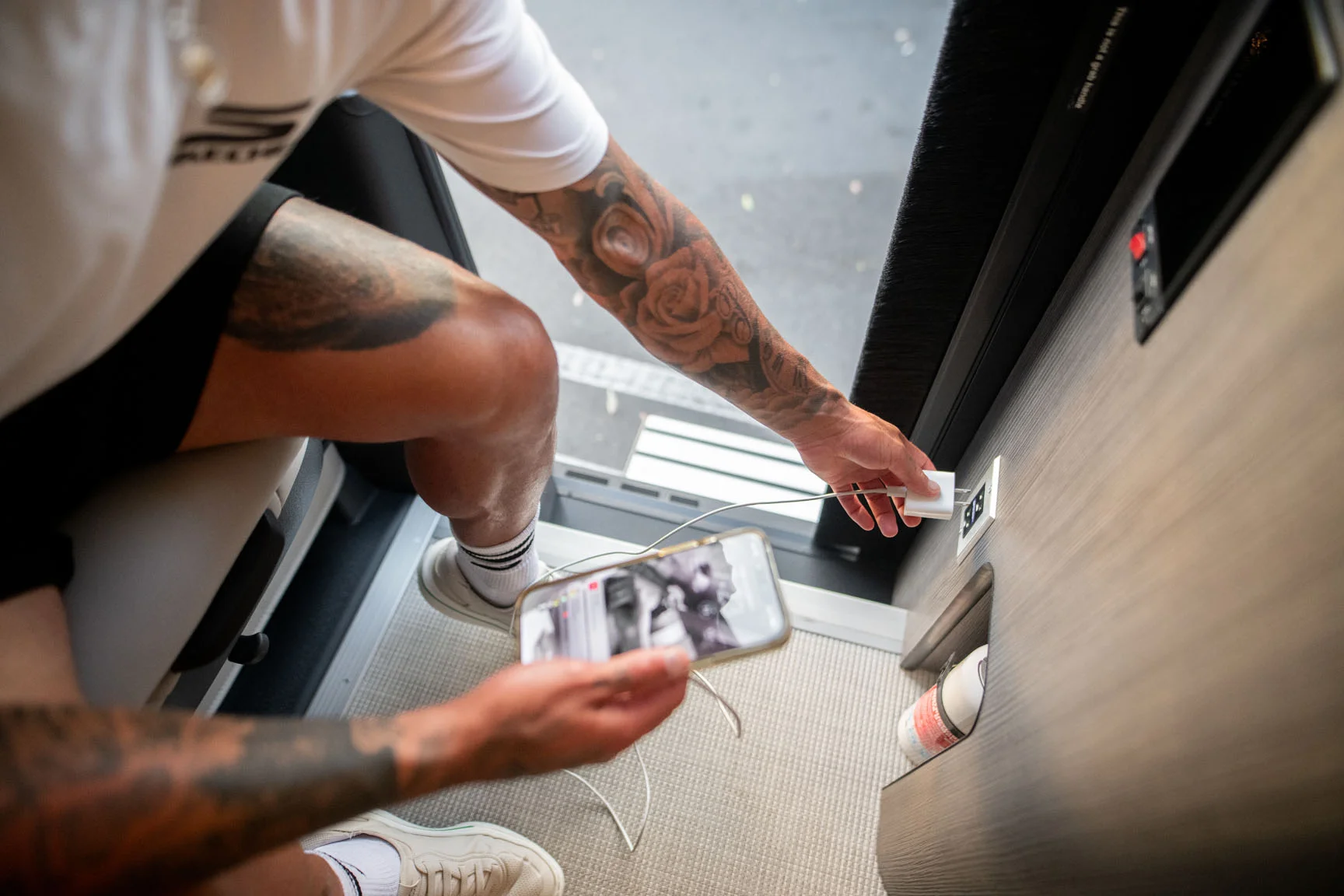 Person with tattoos plugging a smartphone into a wall socket inside a vehicle, wearing white shorts and sneakers.