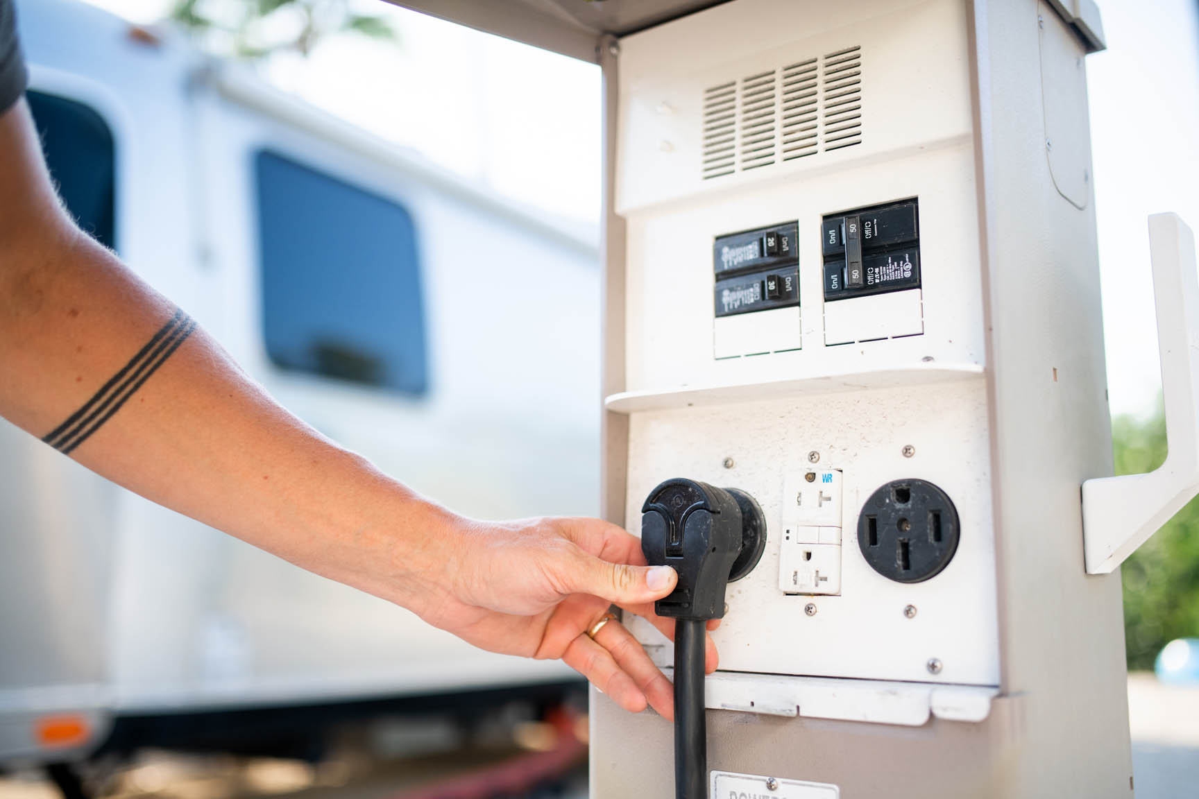 A person’s hand plugs a heavy-duty electrical cord into a power pedestal with multiple outlets at an outdoor RV site. An RV is visible in the blurred background.