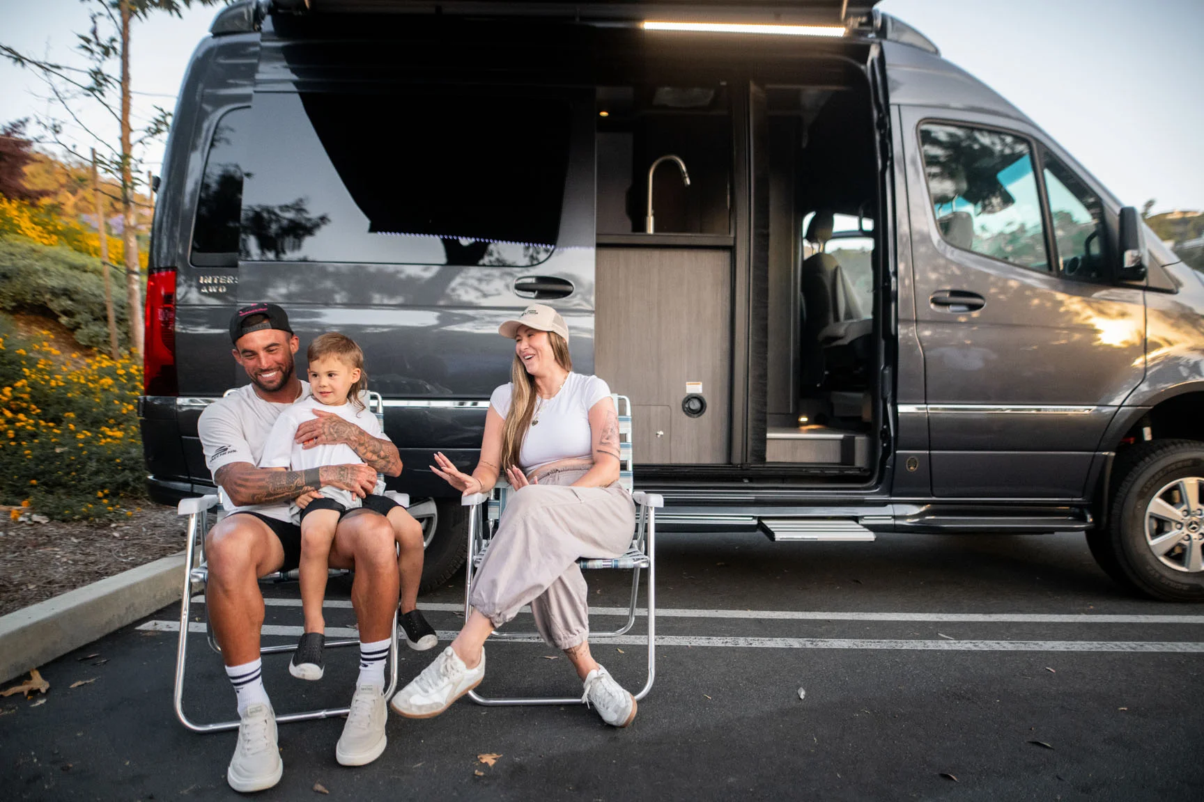 A family of three sits on folding chairs beside a parked camper van, smiling and enjoying time together in a parking lot.