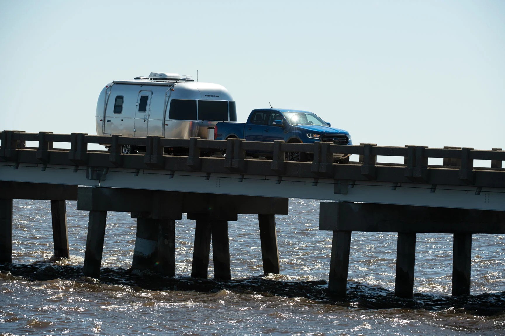 A blue pickup truck towing a silver trailer drives over a bridge above choppy water under a clear blue sky.