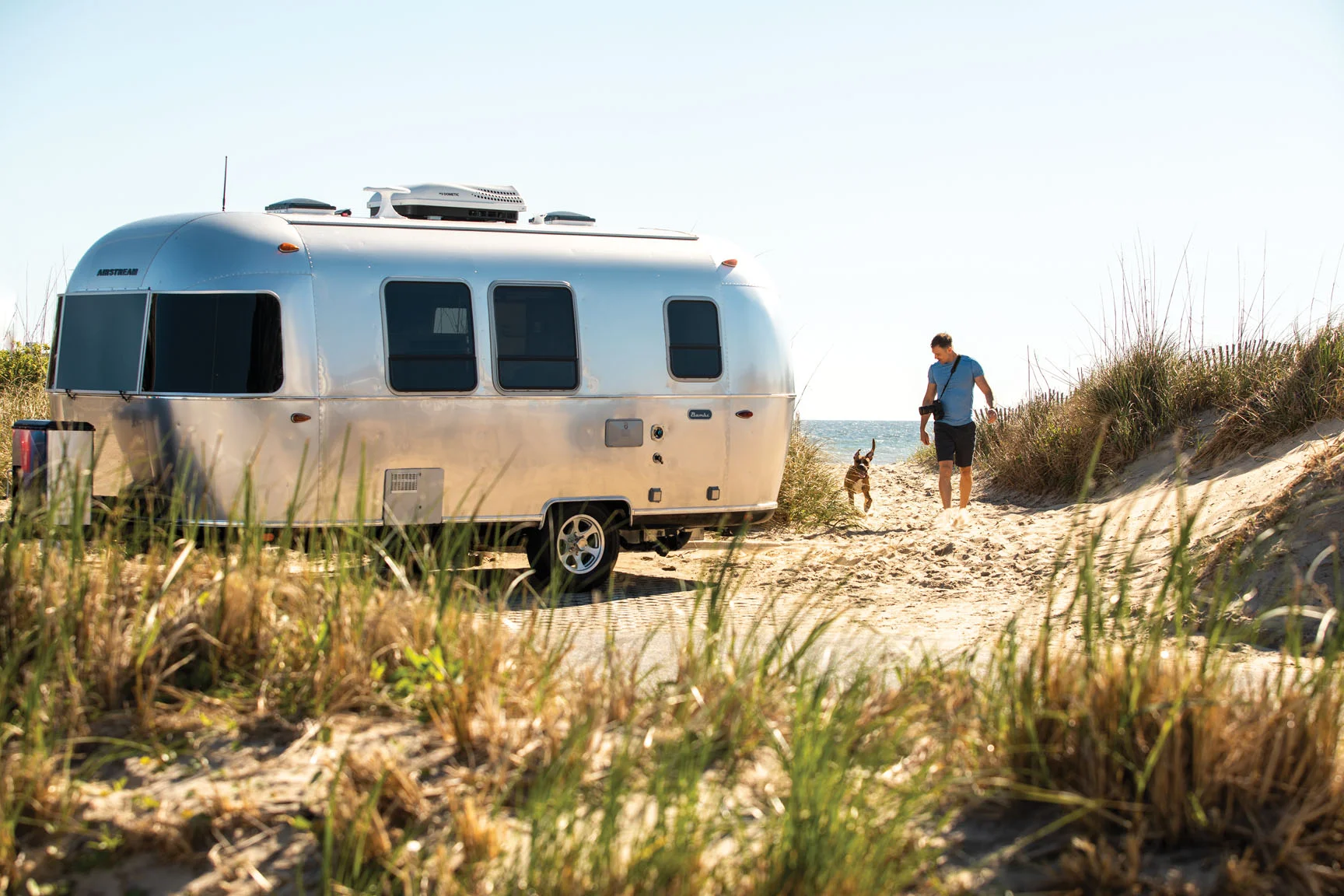 A silver camper trailer parked near a sandy beach with a man walking a dog among grassy dunes under a clear blue sky.