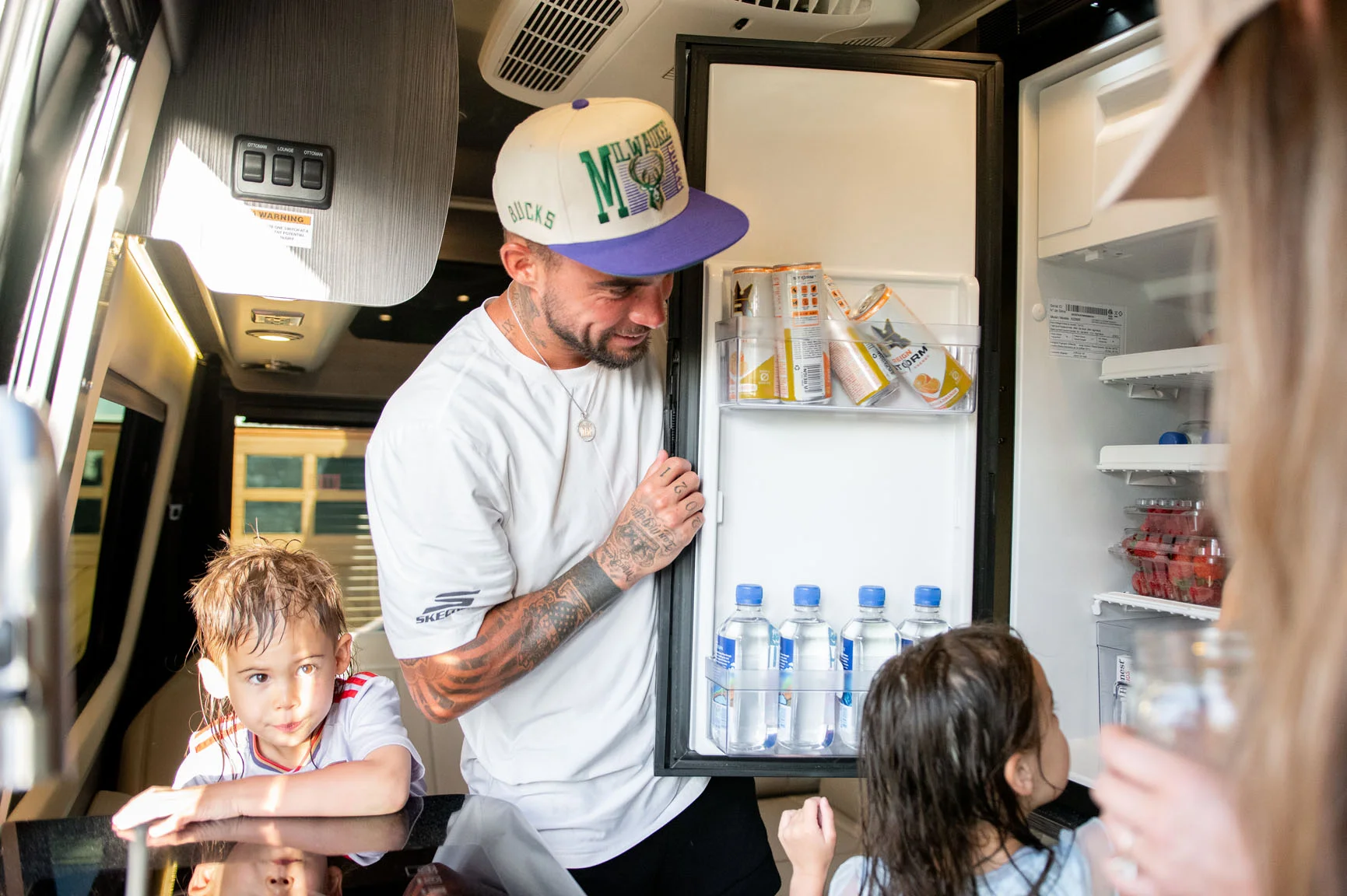A man in a baseball cap opens a fridge in an RV, with two children nearby. The fridge contains drinks and snacks.