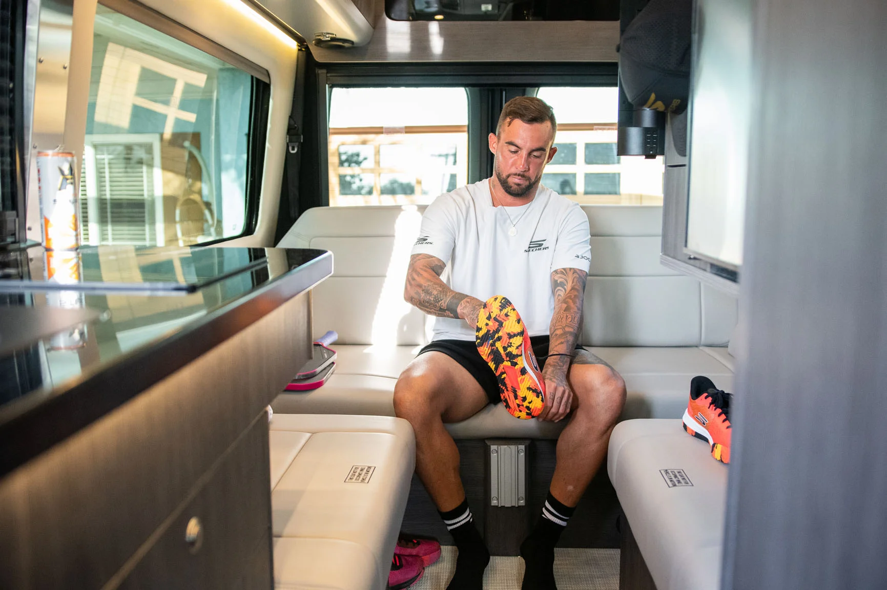 Man sitting in a camper van putting on colorful sports shoes, with tattoos on his arms, wearing a white shirt and black shorts.