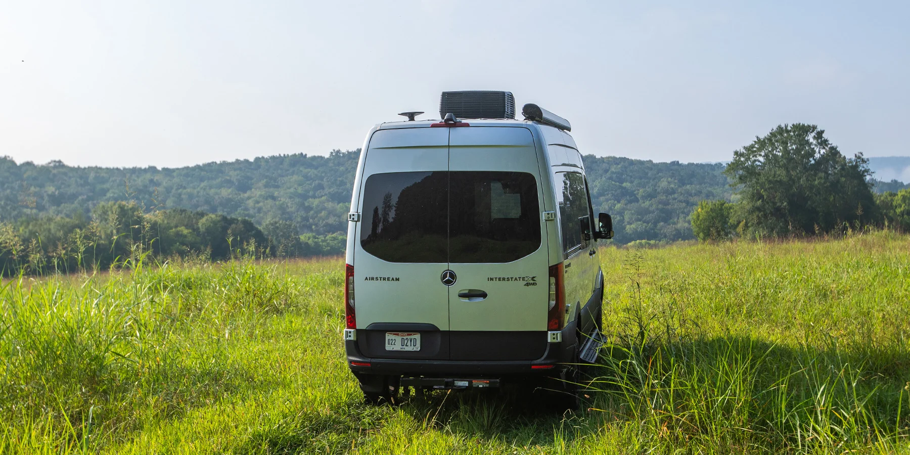 A silver Mercedes-Benz camper van is parked on a grassy field with rolling green hills and trees in the background under a clear sky.