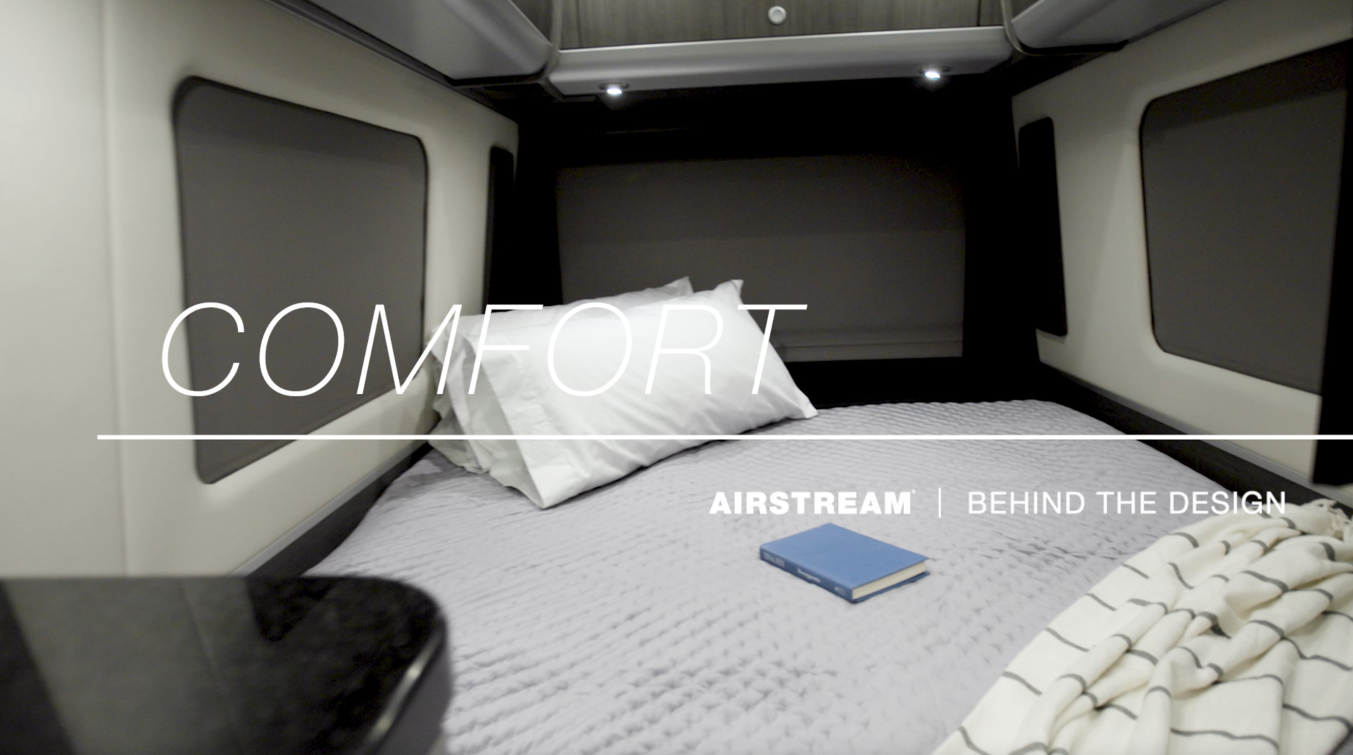 Cozy camper interior with a bed, white pillow, striped blanket, and a book. Text overlay: "COMFORT, AIRSTREAM | BEHIND THE DESIGN."