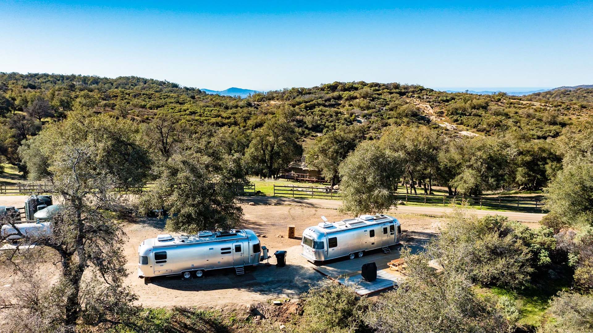 Two silver trailers parked on a spacious, scenic hilltop surrounded by green trees and distant mountains under a clear blue sky.