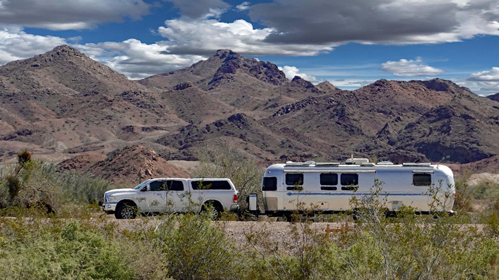 A pickup truck towing an Airstream trailer against a backdrop of rugged desert mountains under a partly cloudy sky.