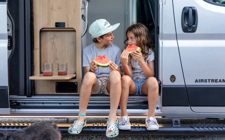 Two children sit in a van doorway, smiling and eating watermelon slices, with drinks on a small shelf nearby. A dog’s head is visible in the foreground.