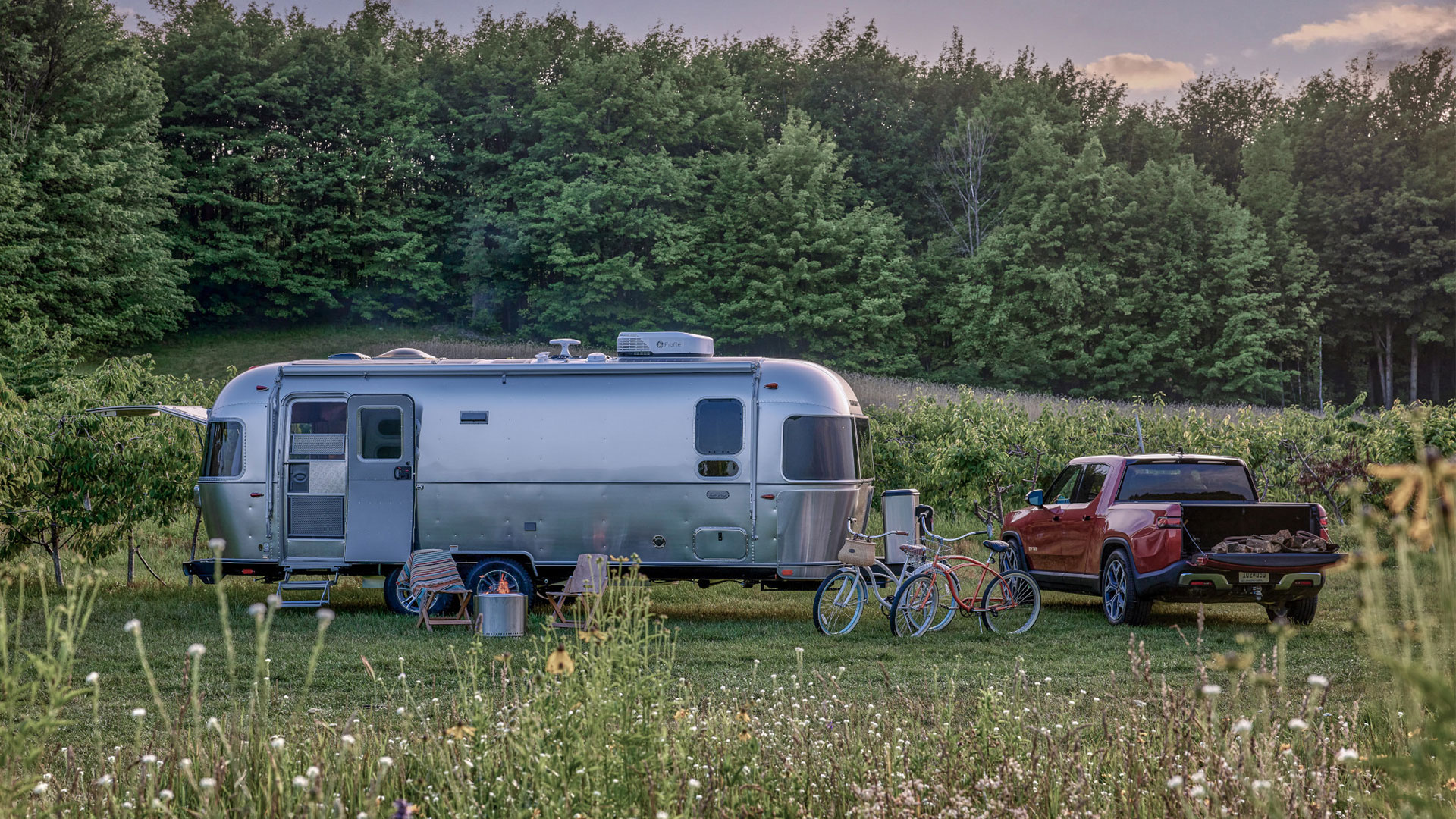 A silver trailer and red truck parked in a grassy field with two bicycles in front, surrounded by lush green trees and wildflowers.
