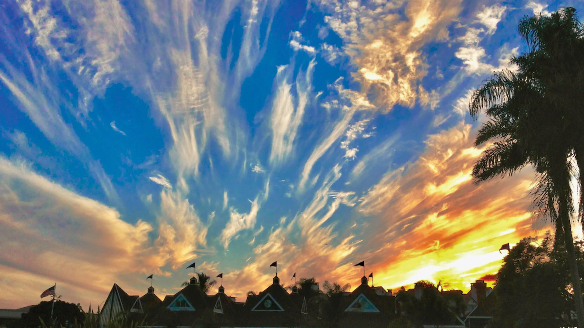 Vivid sunset with wispy clouds streaking across a blue and orange sky above silhouetted houses and palm trees. Flags are visible on rooftops, and the sunlight casts a warm glow over the scene.