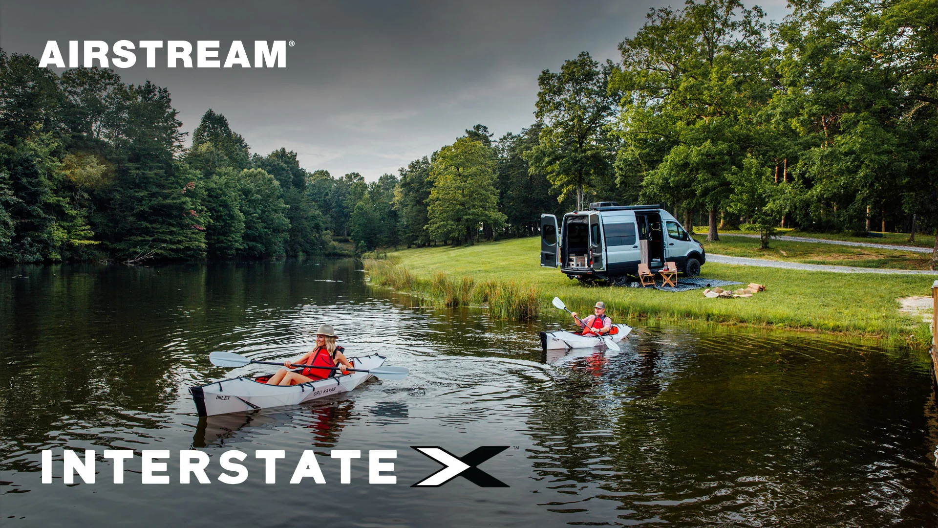 Two people kayaking on a serene lake near an Airstream Interstate X camper van parked on a grassy shore surrounded by trees.
