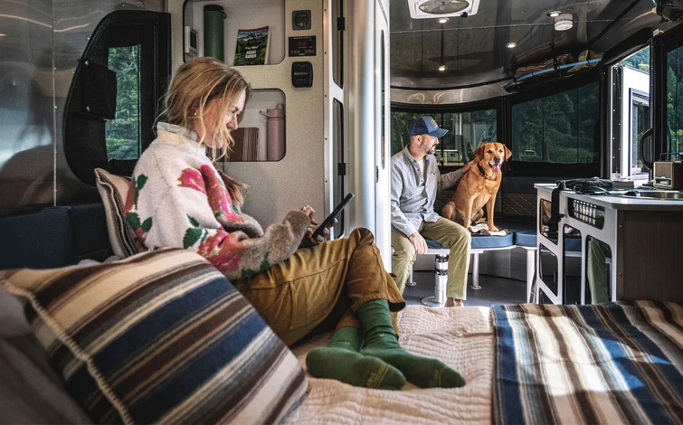 A woman sits on a bed using a tablet in an RV, while a man sits nearby and a brown dog sits on a bench by large windows overlooking trees. The interior looks cozy and well-lit.