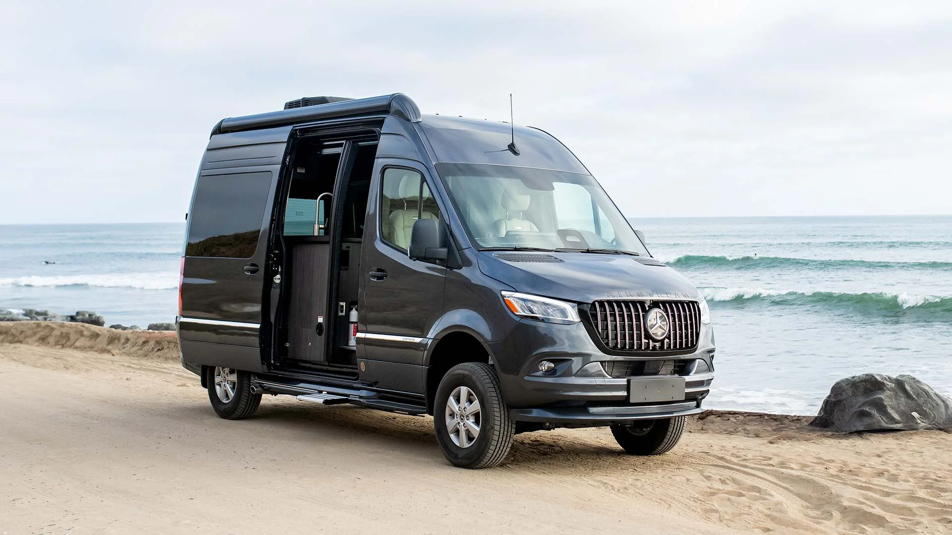 Black camper van parked on a sandy beach with ocean waves in the background and the side door open.