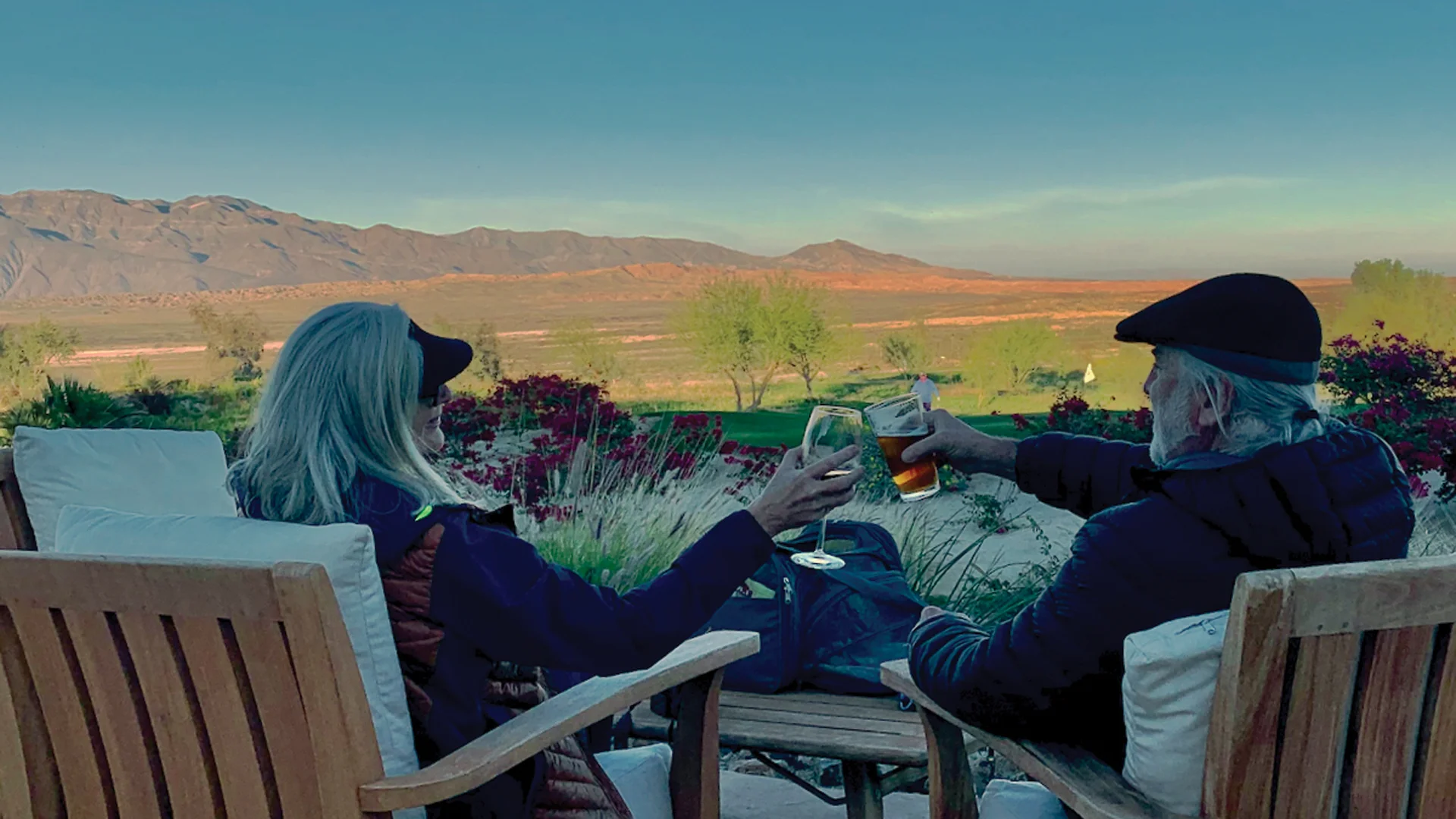 Two people with gray hair sit in wooden chairs outdoors, clinking drinks and facing a scenic landscape with mountains, fields, and a clear blue sky at sunset.