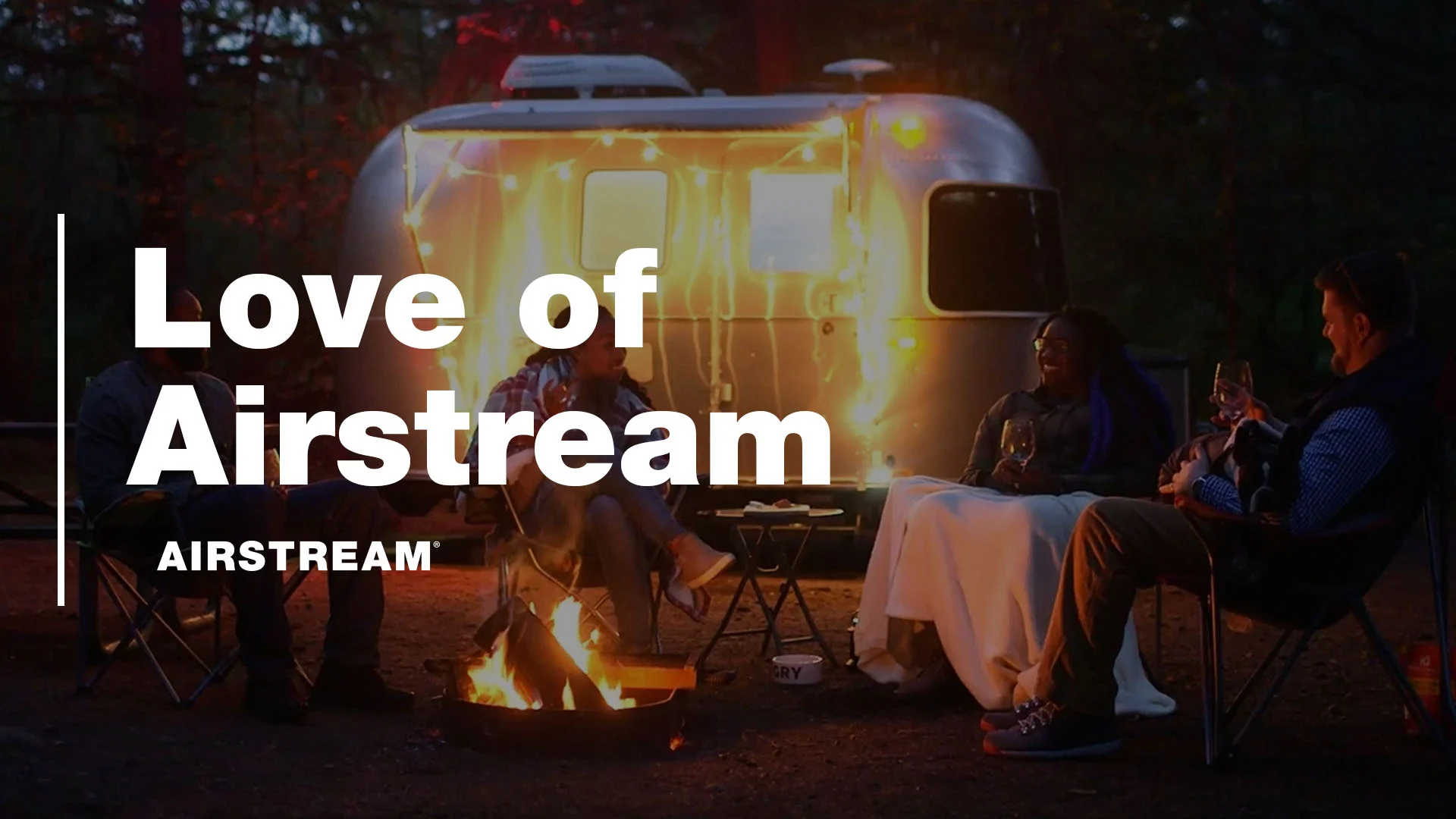 People sitting by a campfire near an Airstream trailer, illuminated by string lights. The text reads "Love of Airstream."