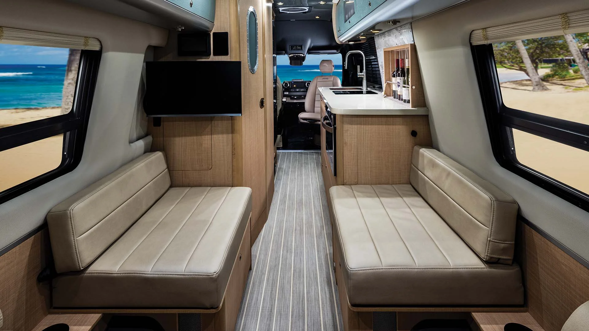 Interior view of a modern camper van with two beige benches, a kitchenette, and windows showing a beach scene.