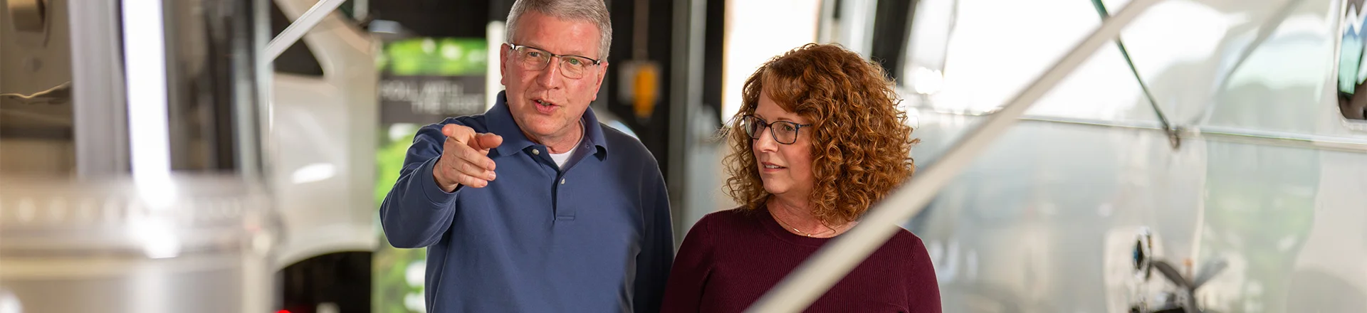 A man and woman walk together in an RV showroom, discussing and pointing towards a modern travel trailer.