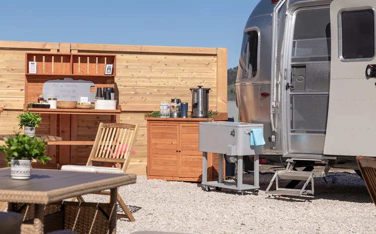 Outdoor kitchenette with wooden cabinets and a silver trailer. Chairs and tables in the foreground, plants for decor, clear blue sky.