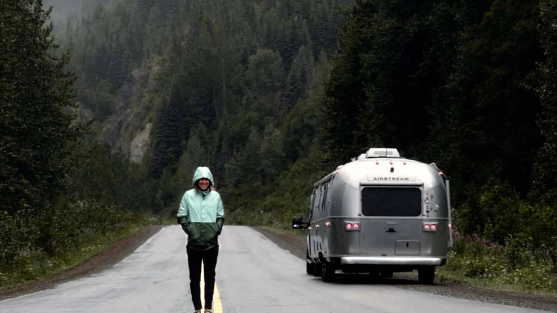 Person in a green jacket walks on a forest road beside a parked silver Airstream trailer, surrounded by dense trees and mountainous landscape.