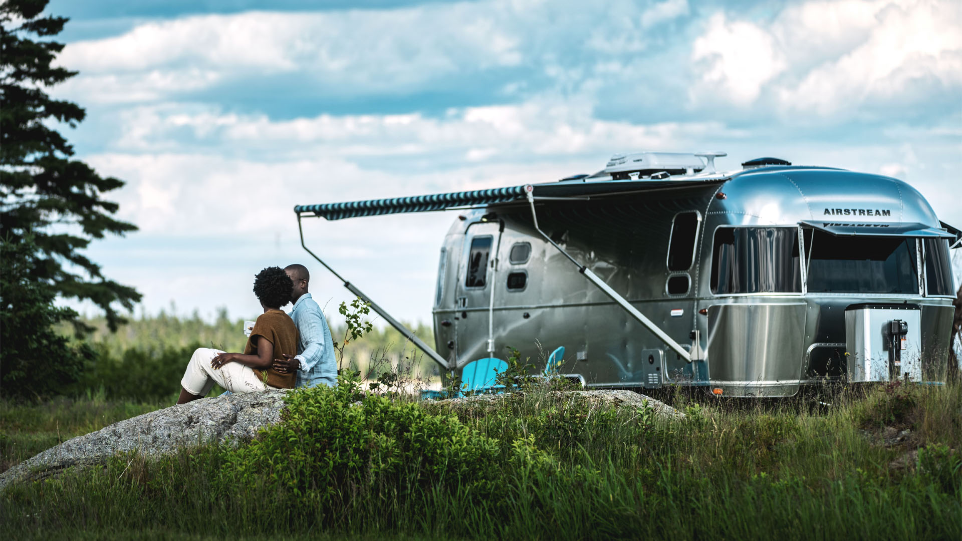 A couple sits together on a rock, facing away, near a modern silver Airstream trailer parked in a grassy area with trees and a cloudy sky in the background.