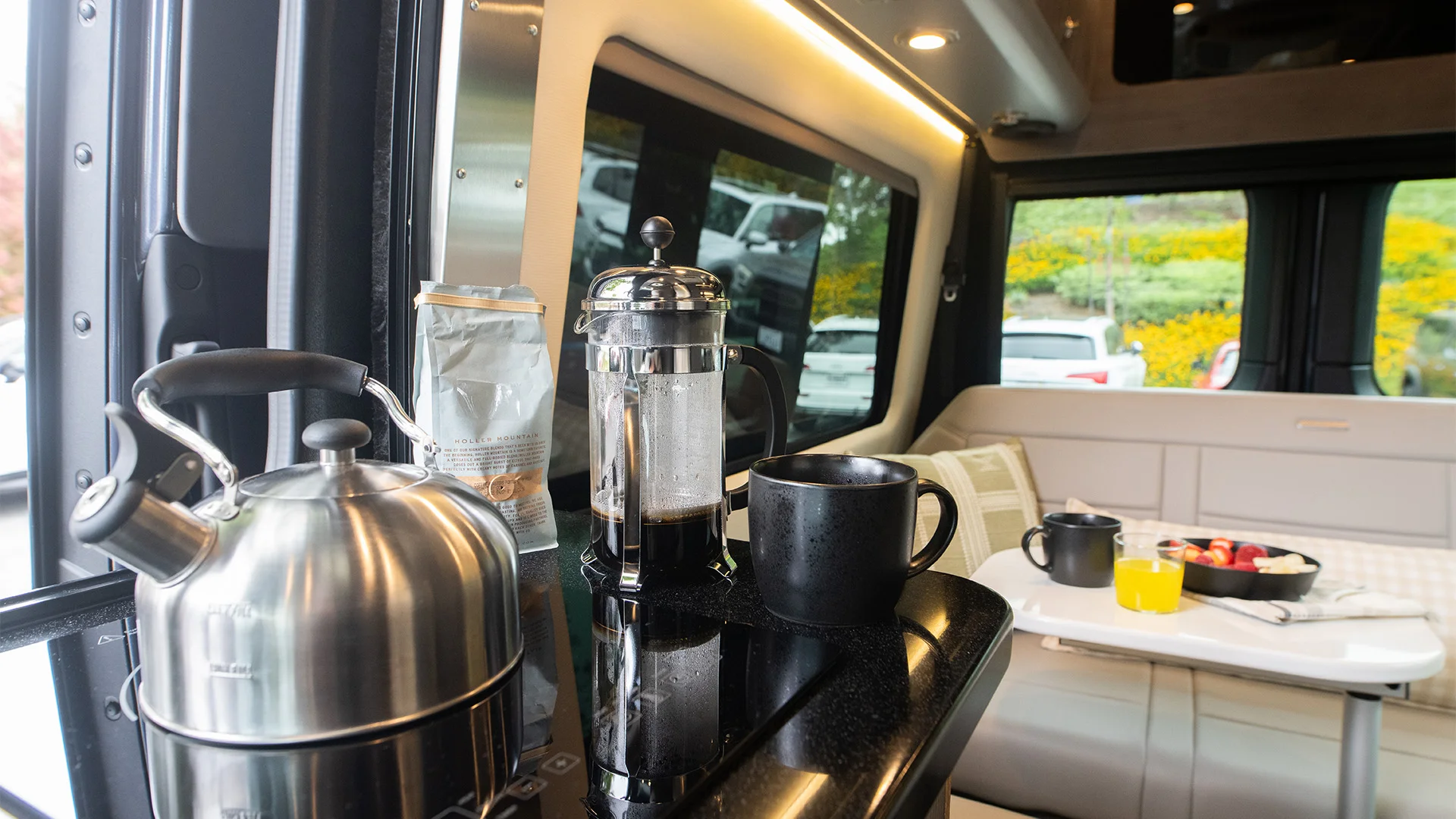 Cozy campervan interior with a stainless steel kettle, French press, and black mugs on a counter. A table set with fruits and yogurt is visible.
