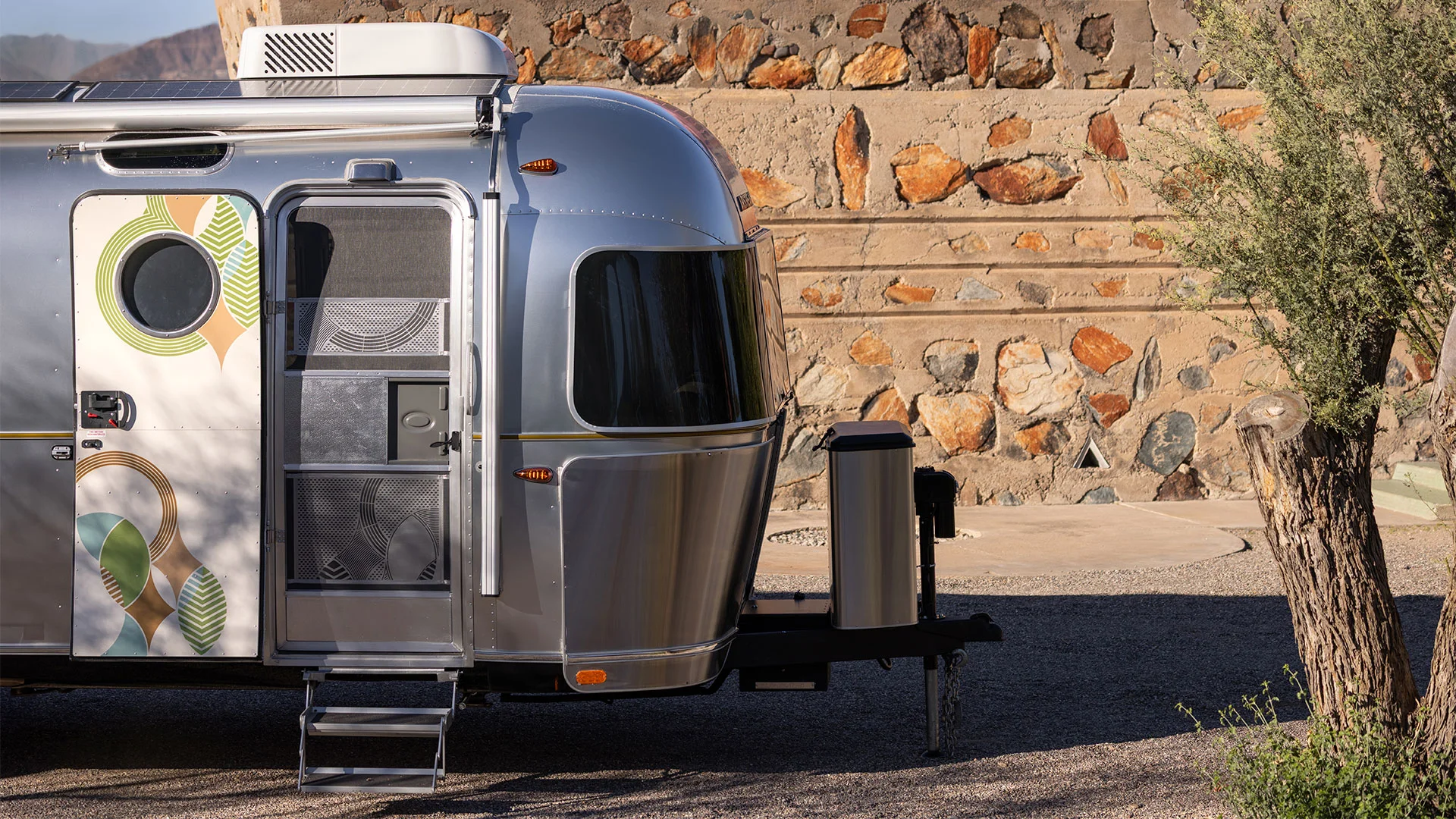 Shiny silver Airstream trailer with patterned door, set against a stone wall and tree in a sunny outdoor setting.