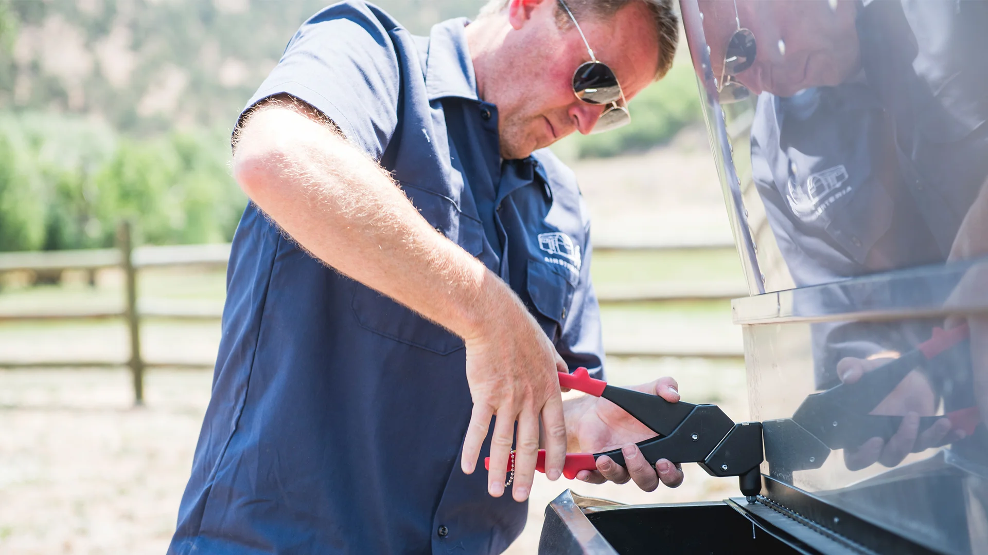 Man in sunglasses and blue shirt using a rivet tool on a metal outdoor structure, with a rural landscape in the background.